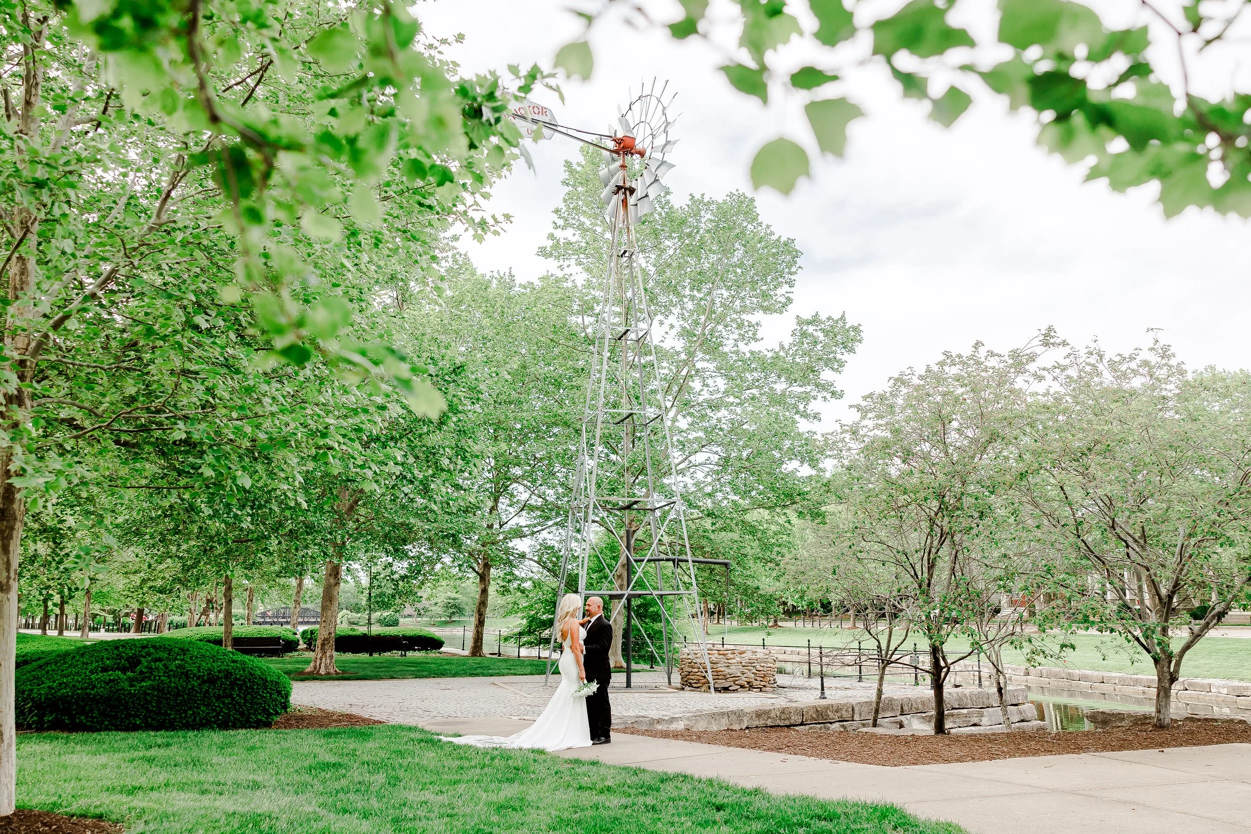 Bride and groom standing together in a park with lush green trees, a windmill, and a small pond in the background on a clear day.