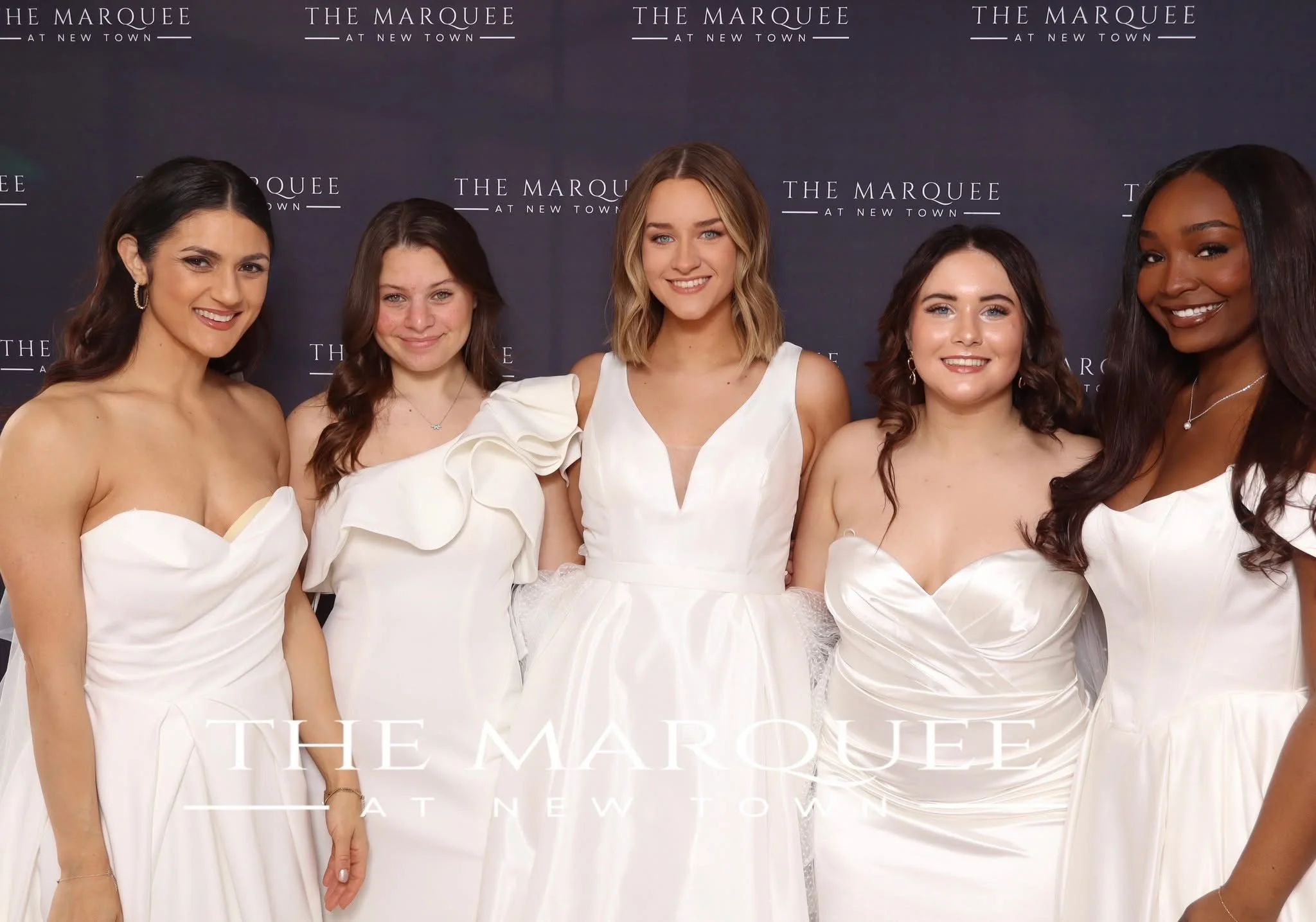 five women all wearing different white dresses. they're modeling at the marquee. there's a backdrop behind them saying the marquee. they're all glowing and smiling so radiantly. 