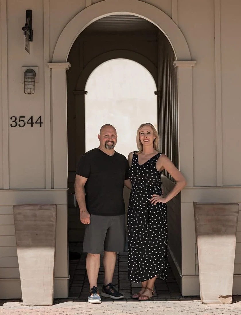 A man and a woman, owners of The Marquee at New Town, standing outside the event tent with a large arched doorway. The man is wearing a black t-shirt and gray shorts, while the woman is dressed in a black and white polka dot dress. Both are smiling.