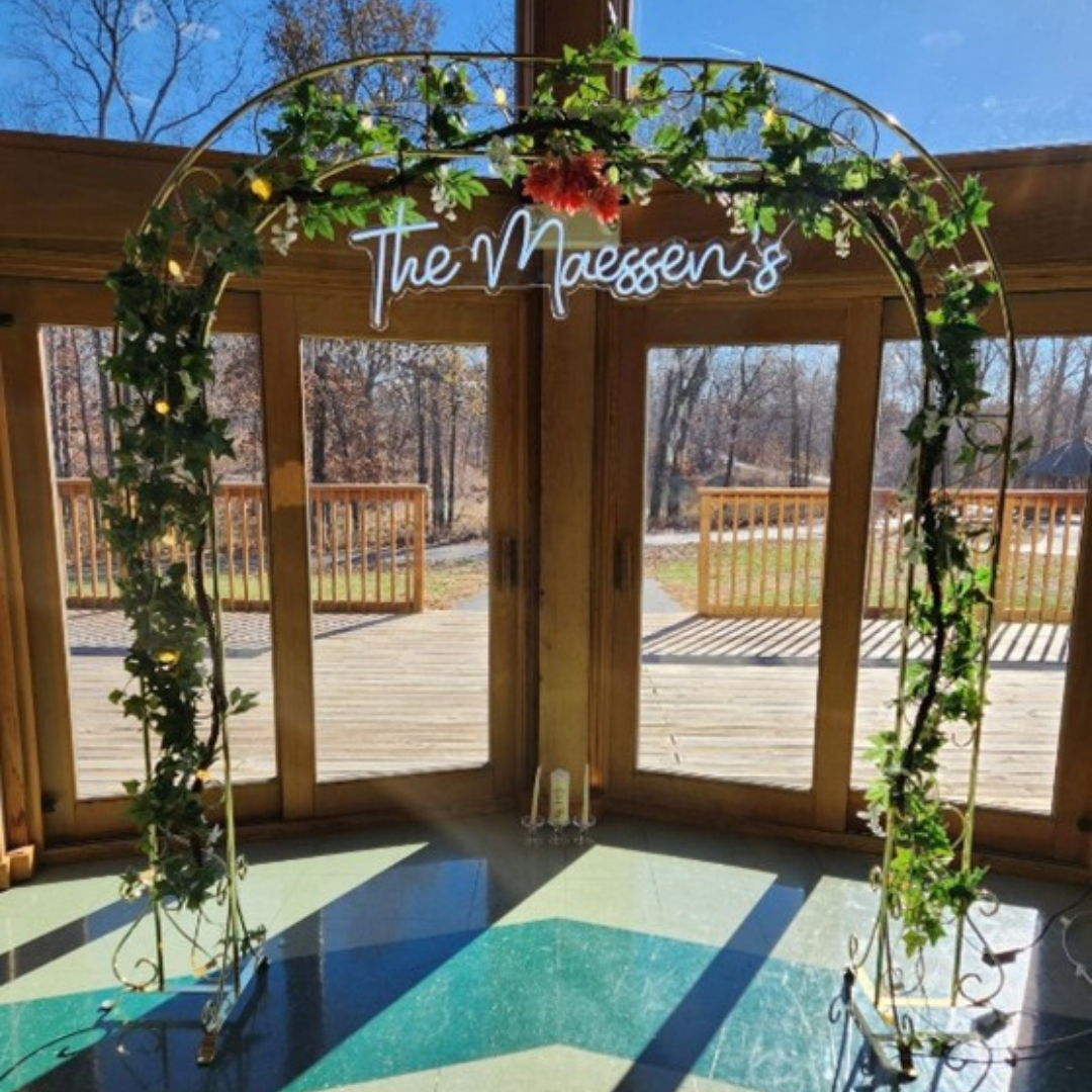 Decorated gold arch with green leaves and flowers and a lit neon sign that reads "The Maassen's" inside a wooden house with large windows and a deck outside available for rental at The Marquee at New Town.