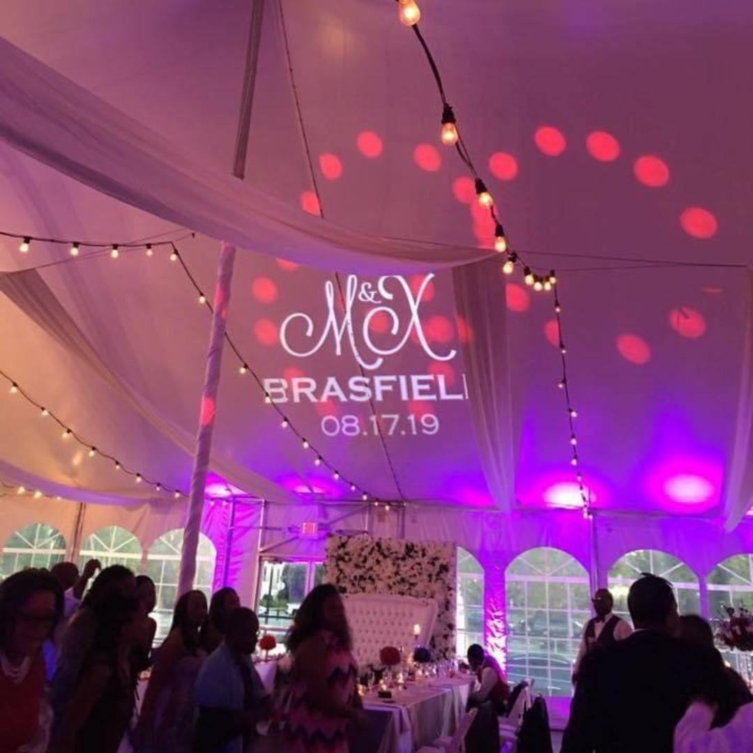 Decorative event tent with string lights, pink and purple lighting, and a floral backdrop. Projected onto the ceiling is a wedding monogram with the names Max & Mac, the last name Brasfield, and the date August 17, 2019. Guests are seated at tables, with some standing and socializing.