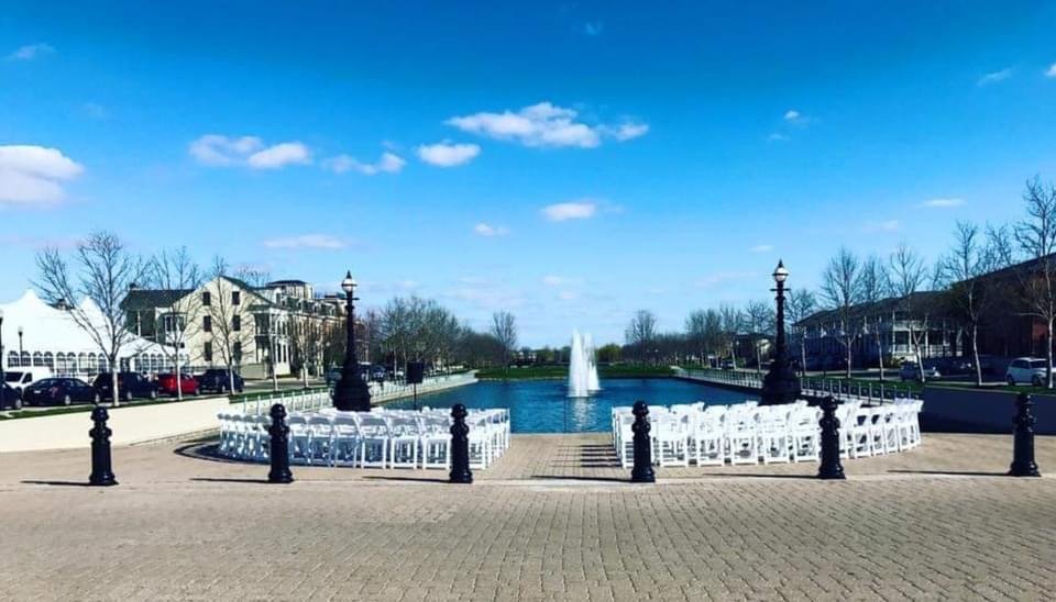 Outdoor scene with a fountain in the middle of a pond, surrounded by white chairs arranged for an event, with a paved area, vintage street lamps, residential buildings and leafless trees in the background, under a blue sky with some clouds.
