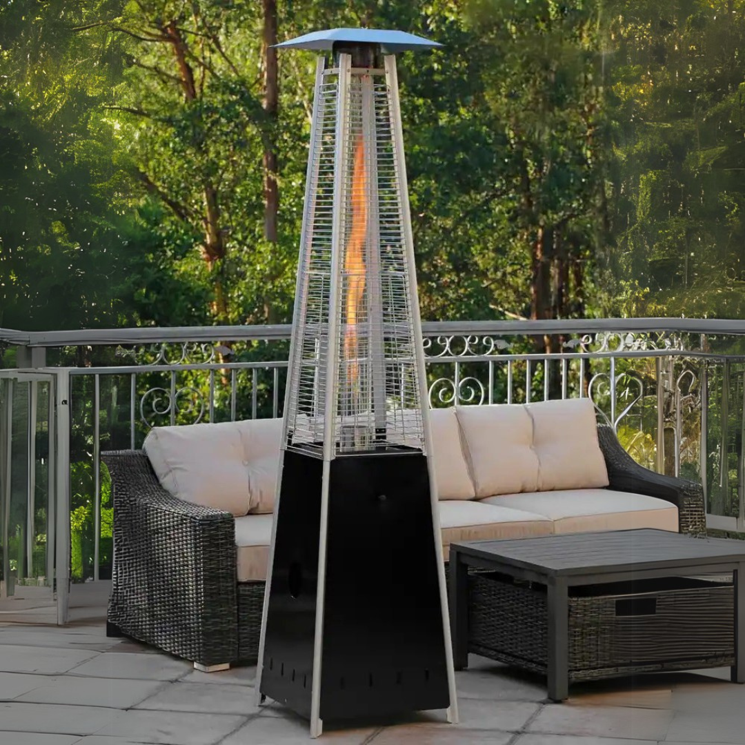 Outdoor patio setup with a wicker sofa, a rectangular coffee table, and a tall, pyramid-shaped outdoor heater on a tiled deck, surrounded by a white metal railing and lush green trees in the background.