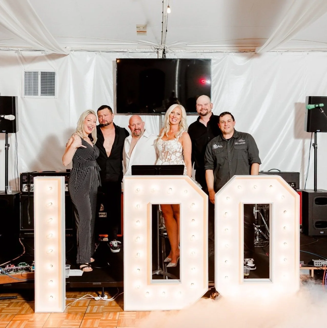 Group of six people at a wedding, standing behind illuminated large letters spelling 'I DO', for rent at The Marquee at New Town for events and weddings, with a white backdrop and equipment around.