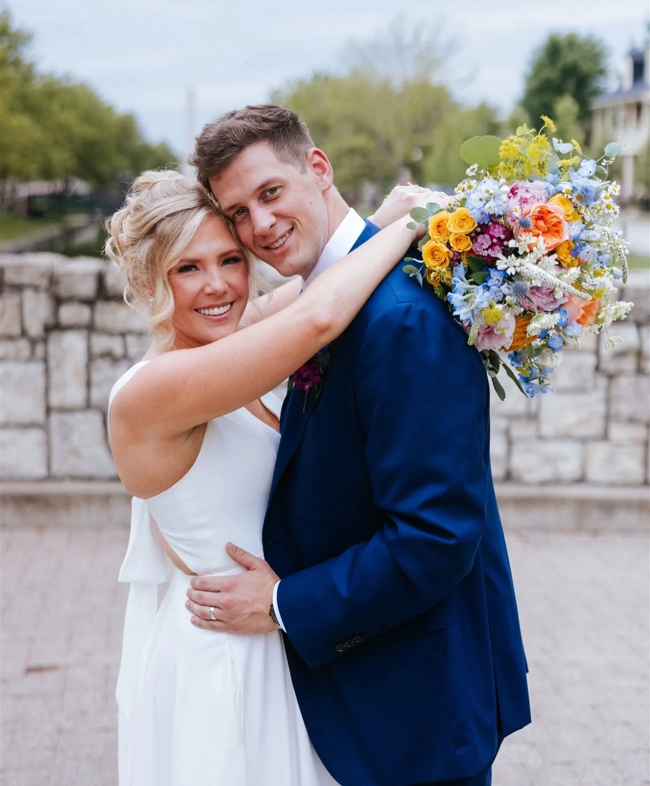 A bride and groom taking pictures on a beautiful bridge overlooking New Town St. Charles canals. She is wearing a white dress and he is wearing a blue suit and she is holding a bouquet of very colorful flowers.