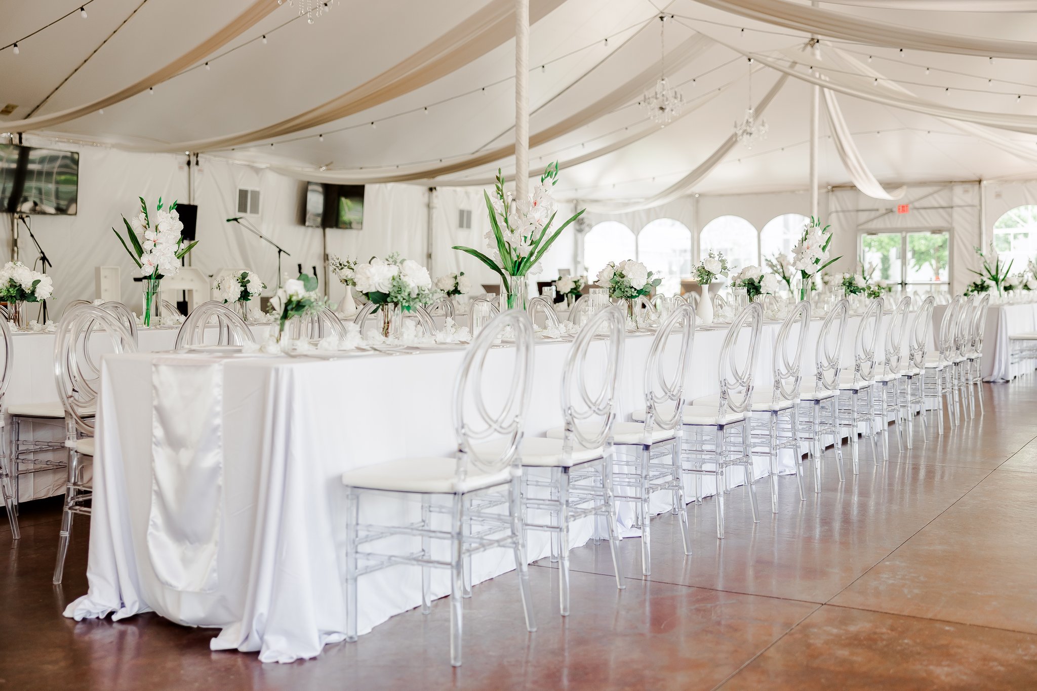 white event tent with tables set up for table reception, long white table with white chairs flower centerpieces, and tan fabric draped from the ceiling, and string lights. 