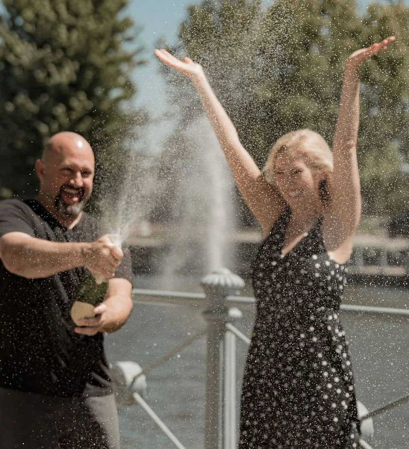 A man and a woman celebrating by popping a champagne bottle, with champagne spray in the air, outside The Marquee at New Town event tent on closing day overlooking New Town canals with fountains.