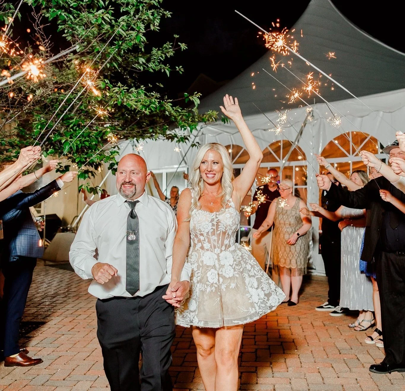 A woman in a floral dress and a man in a white shirt with a black tie walking hand in hand through a line of guests holding sparklers during a wedding reception send-off at night.