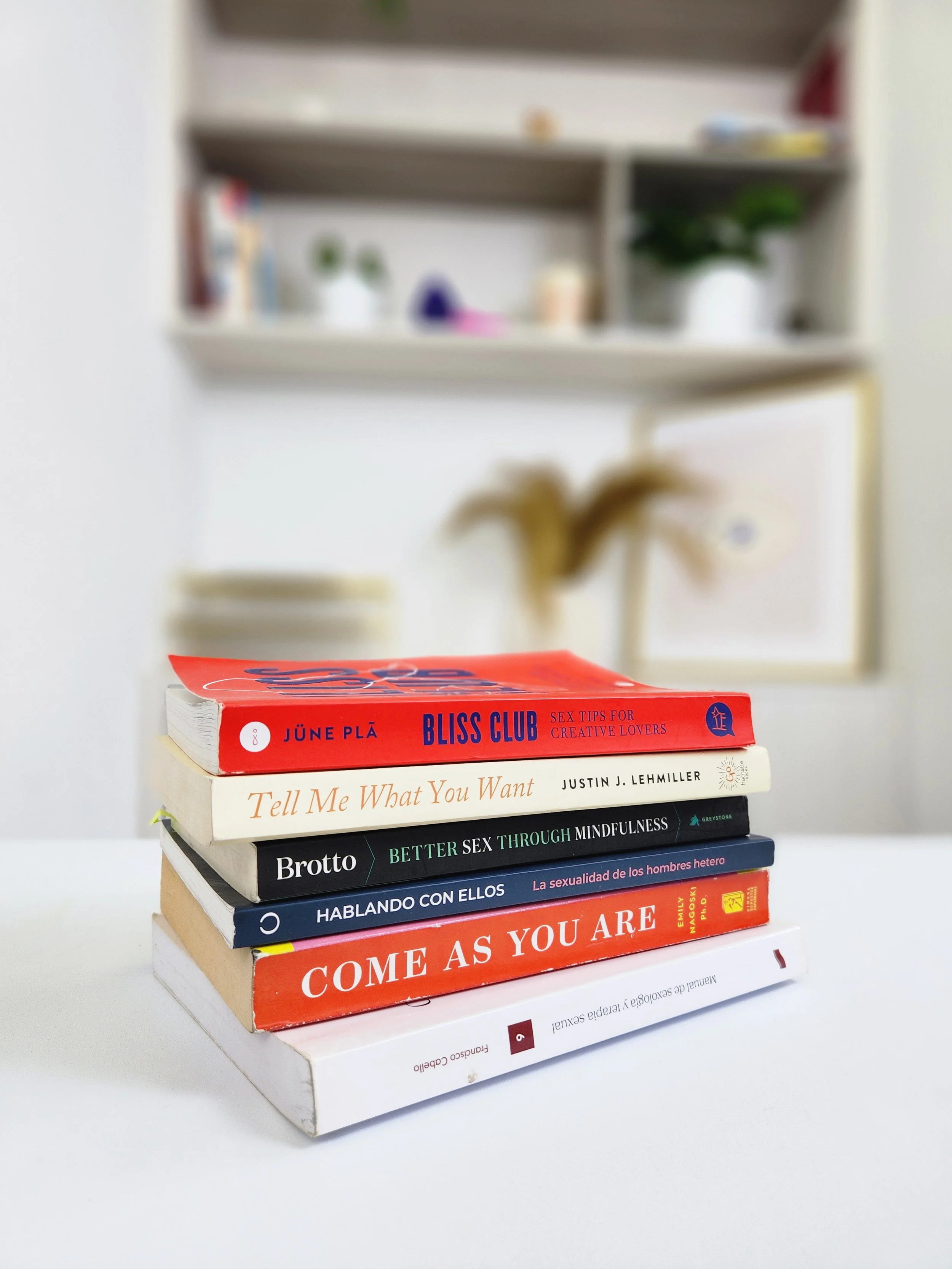 Un conjunto de seis libros apilados sobre una mesa blanca. Los libros están en inglés y español, y tratan sobre temas de sexualidad y relaciones. Al fondo, se ve una estantería borrosa con plantas y decoraciones.
