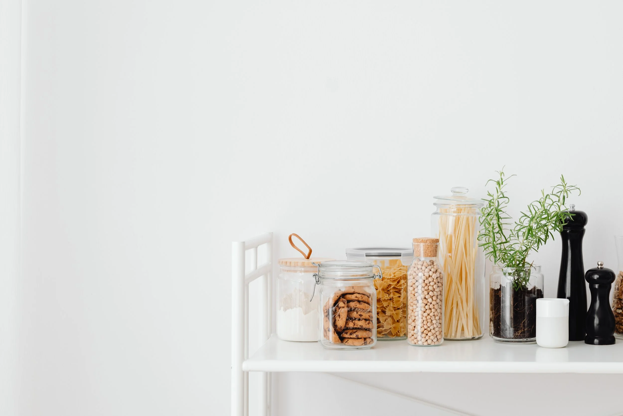 Various jars and containers of dry pasta, cookies, and spices on a white kitchen table, with fresh herbs and black pepper shakers in the background.