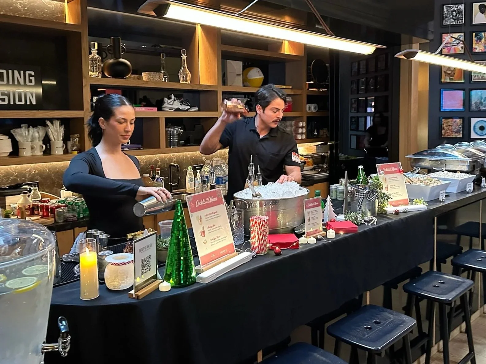 Two people behind a wooden bar with a sign that reads 'PRETTYLITTLETHING,' making drinks outdoors on a sunny day with trees and palm trees in the background. One person is pouring a drink, and the other is shaking a cocktail shaker.