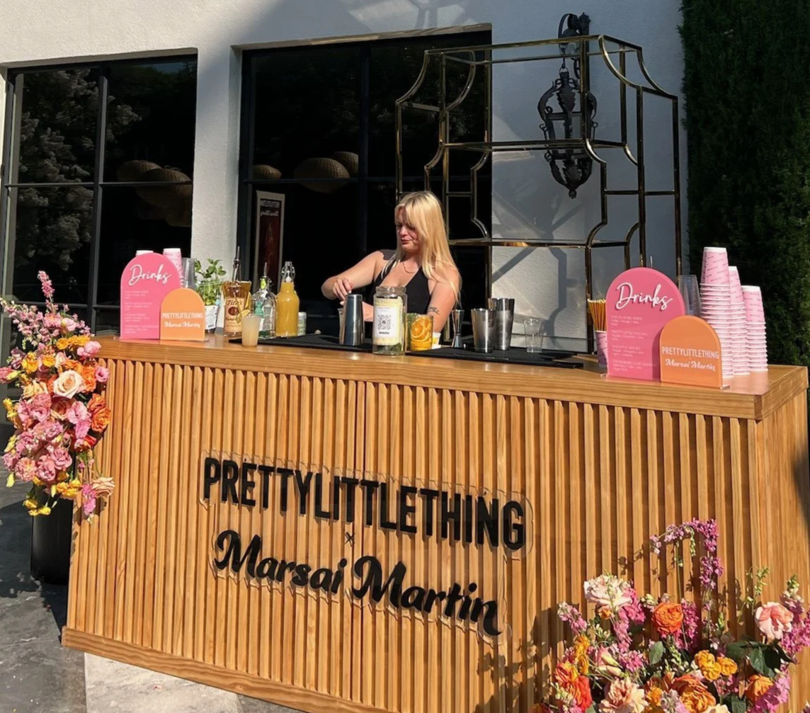 A woman behind a wooden outdoor bar with pink flowers and drink signs, with the words 'PRETTY LITTLE THING Marsai Martin' on the front.