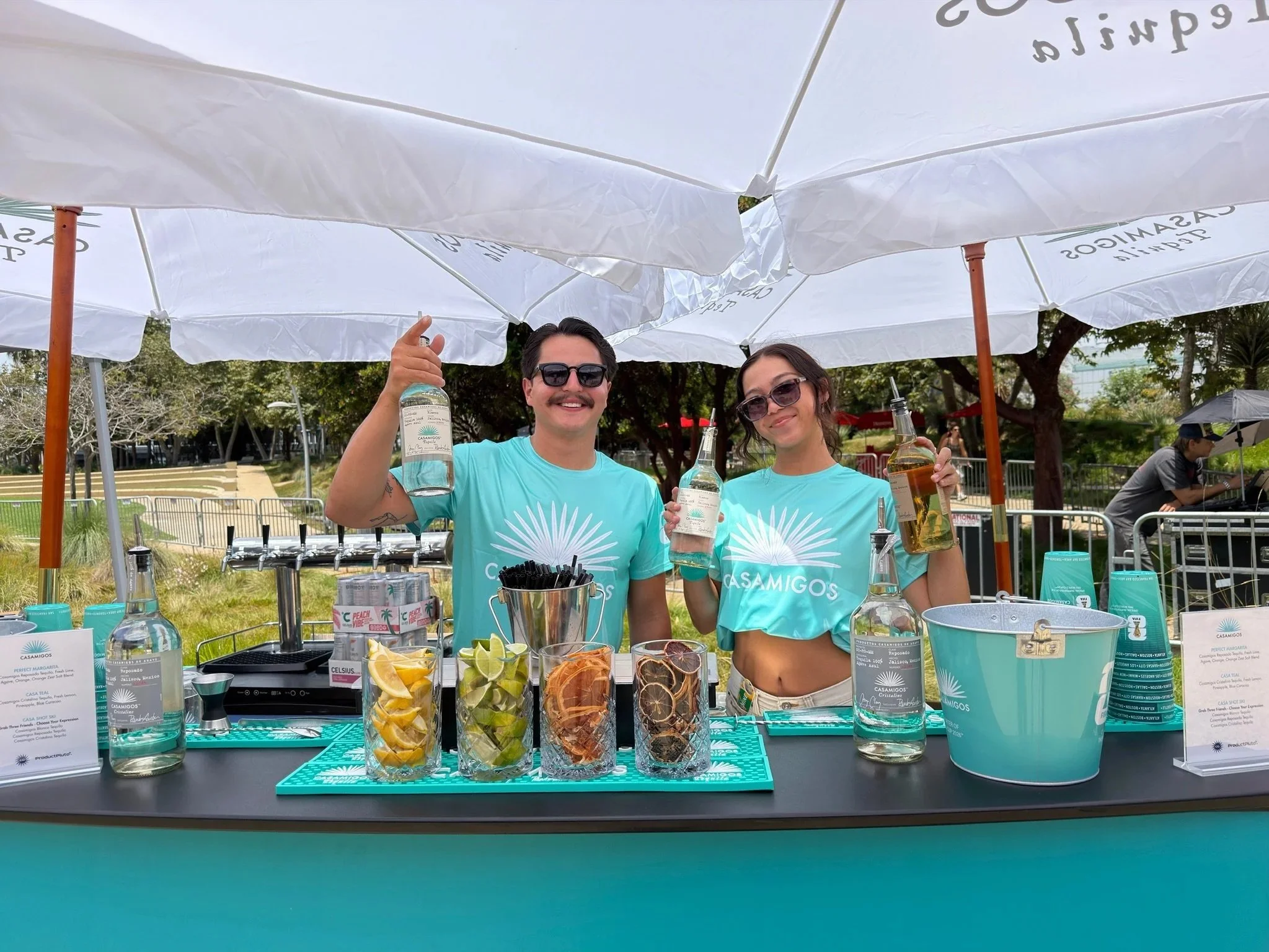 Two people behind a wooden bar with a sign that reads 'PRETTYLITTLETHING,' making drinks outdoors on a sunny day with trees and palm trees in the background. One person is pouring a drink, and the other is shaking a cocktail shaker.