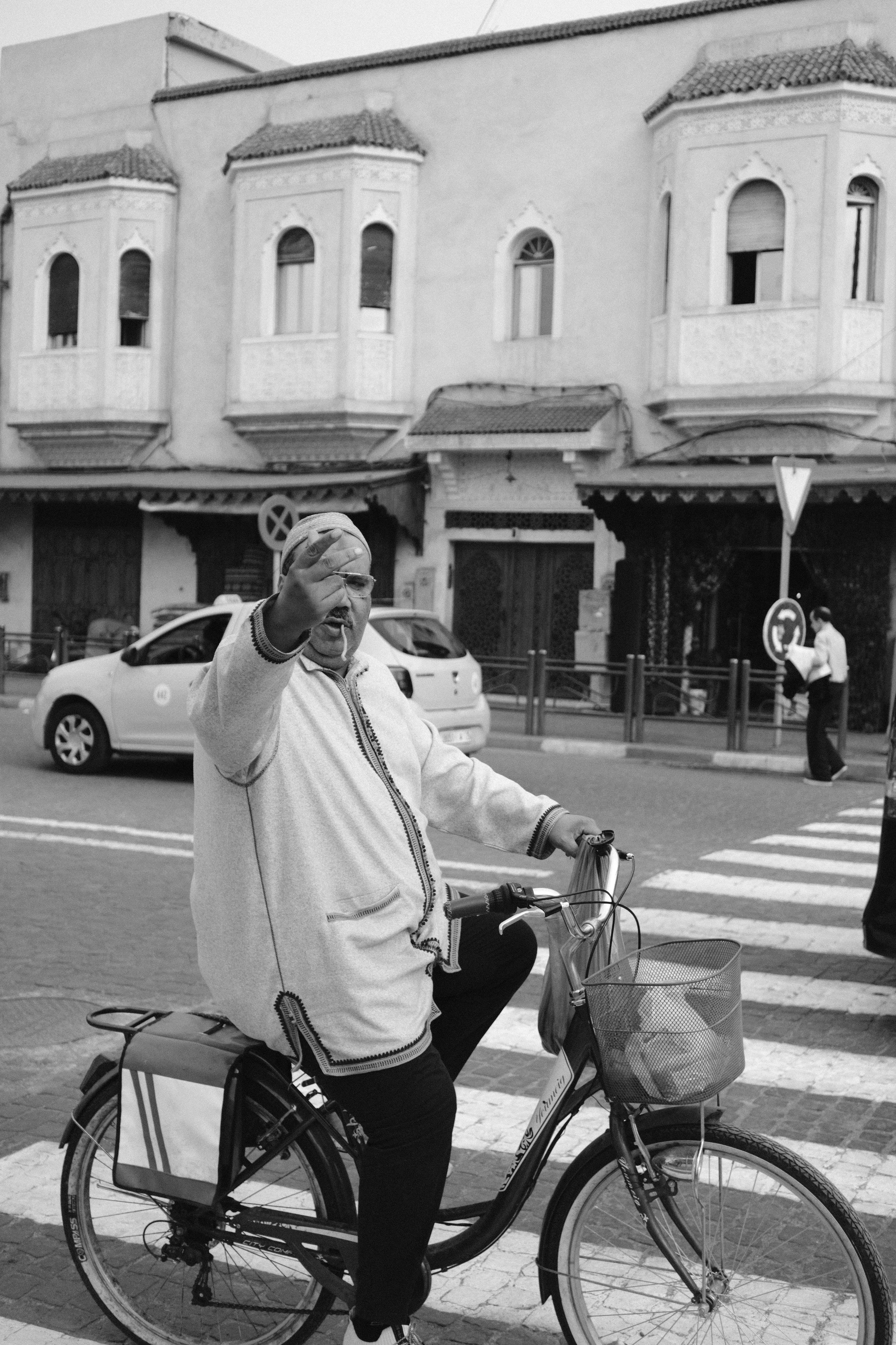 Black and white portrait of a woman on a bicycle on a town street, Taghazout, Morocco - fine art print by Zyan Sachs