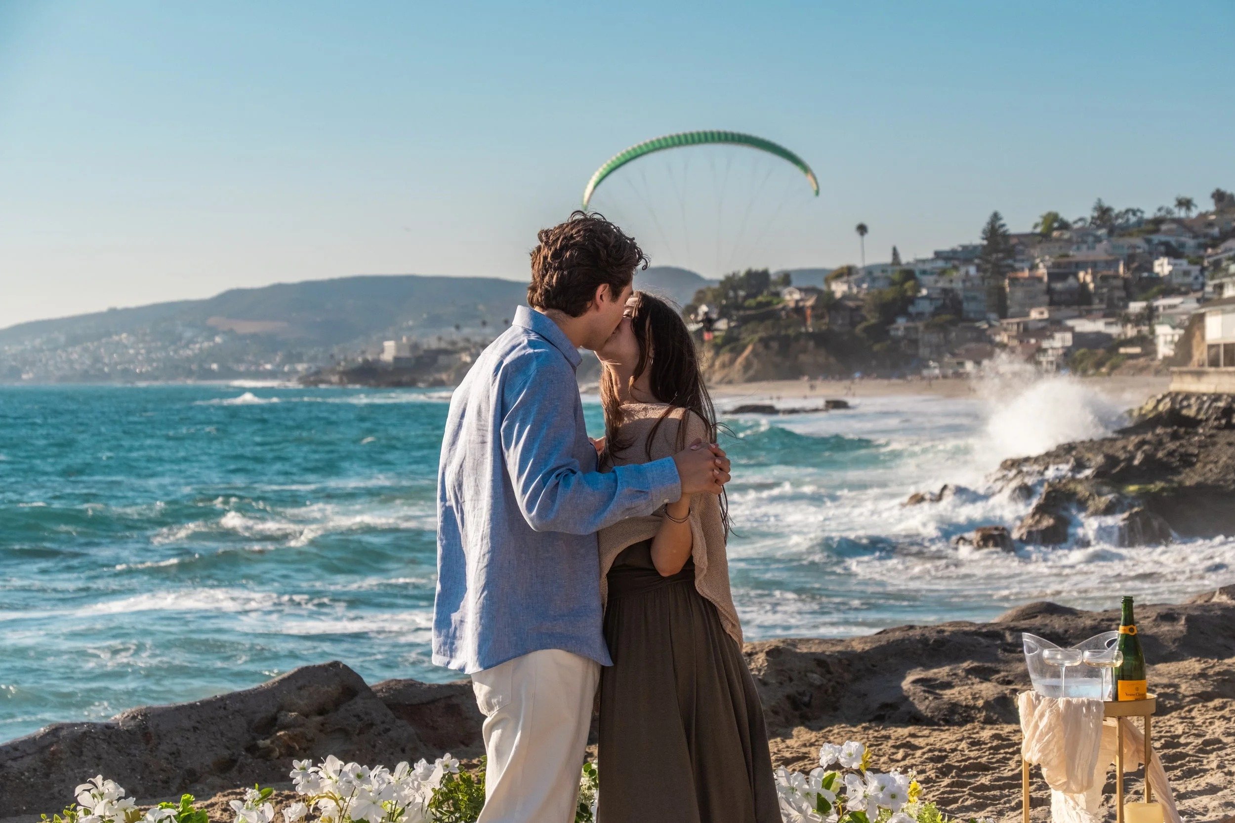 Couple kissing on the rocks after a beach proposal with a paraglider overhead, Southern California - proposal photography by Zyan Sachs