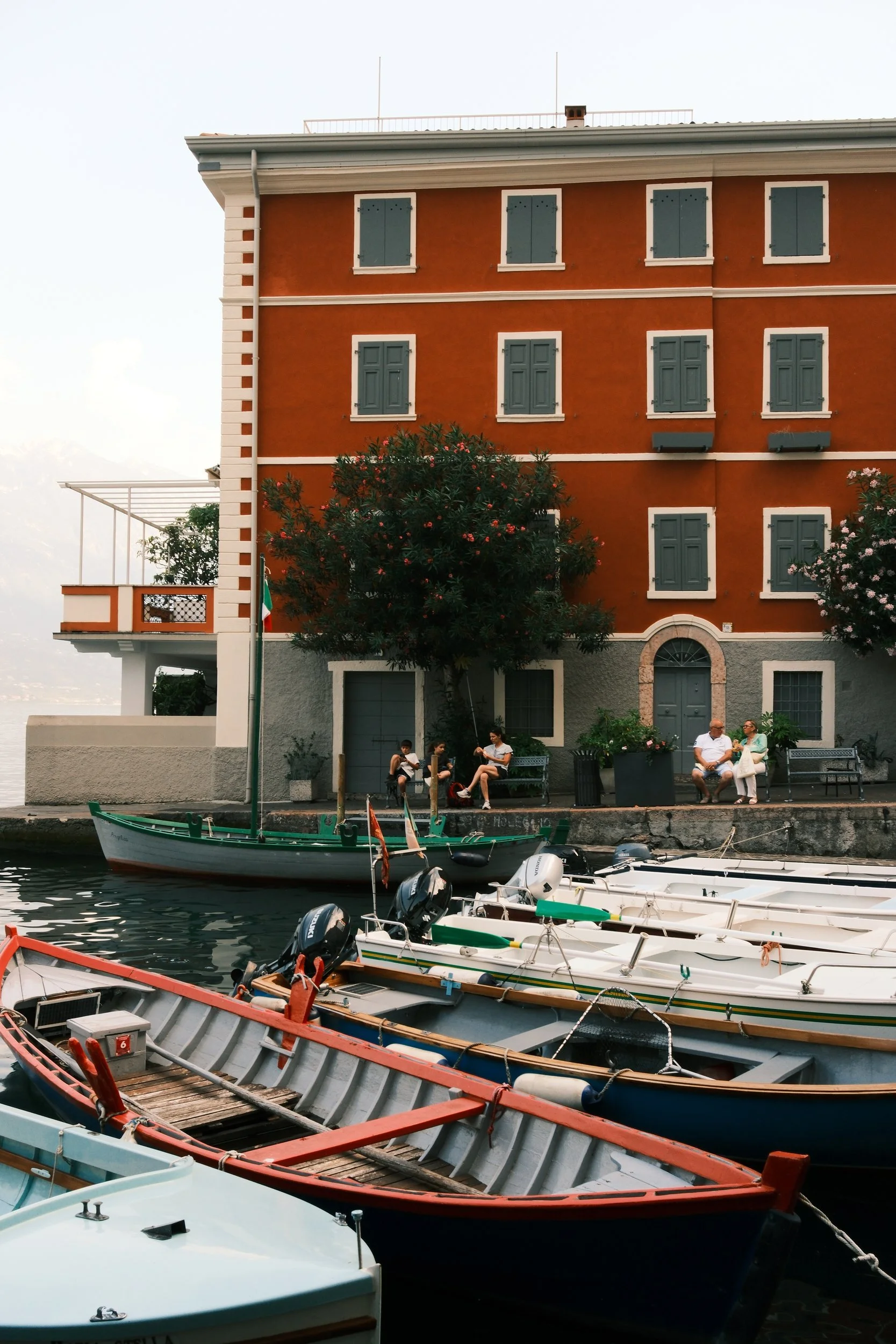 Red-orange lakeside building with green shutters above moored rowboats in Limone sul Garda, Lake Garda, Italy, by Zyan Sachs