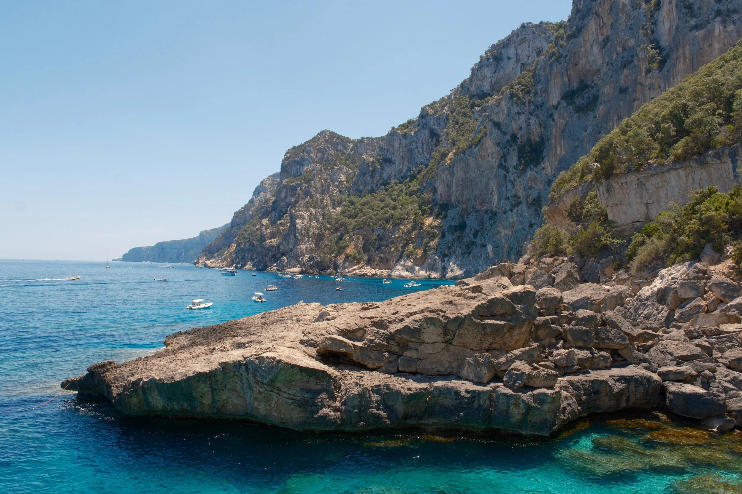 Rocky ledge and towering limestone cliffs with boats anchored along the coast, Gulf of Orosei, Sardinia, Italy, by Zyan Sachs