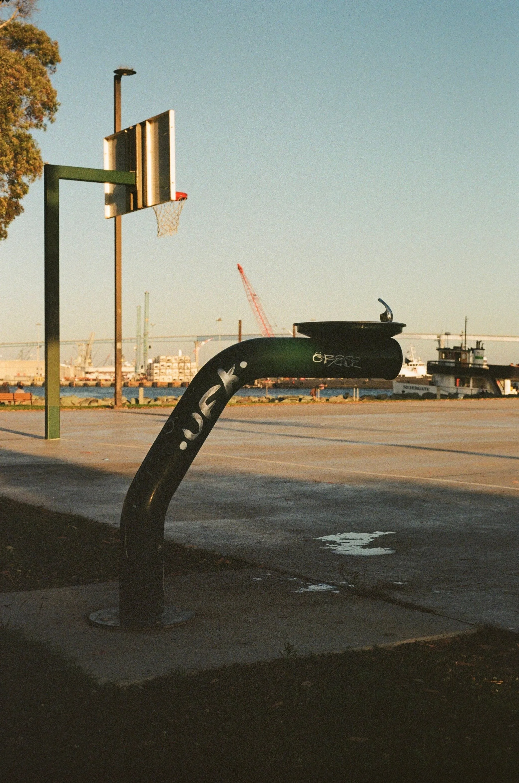 Basketball hoop and drinking fountain by the bay with shipyard cranes, San Diego, California - fine art print by Zyan Sachs