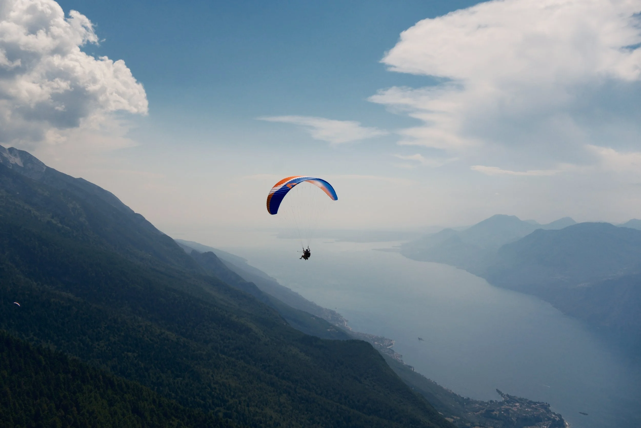 Paraglider with an orange and blue wing soaring above the forested slopes of Monte Baldo over Lake Garda, Italy, by Zyan Sachs