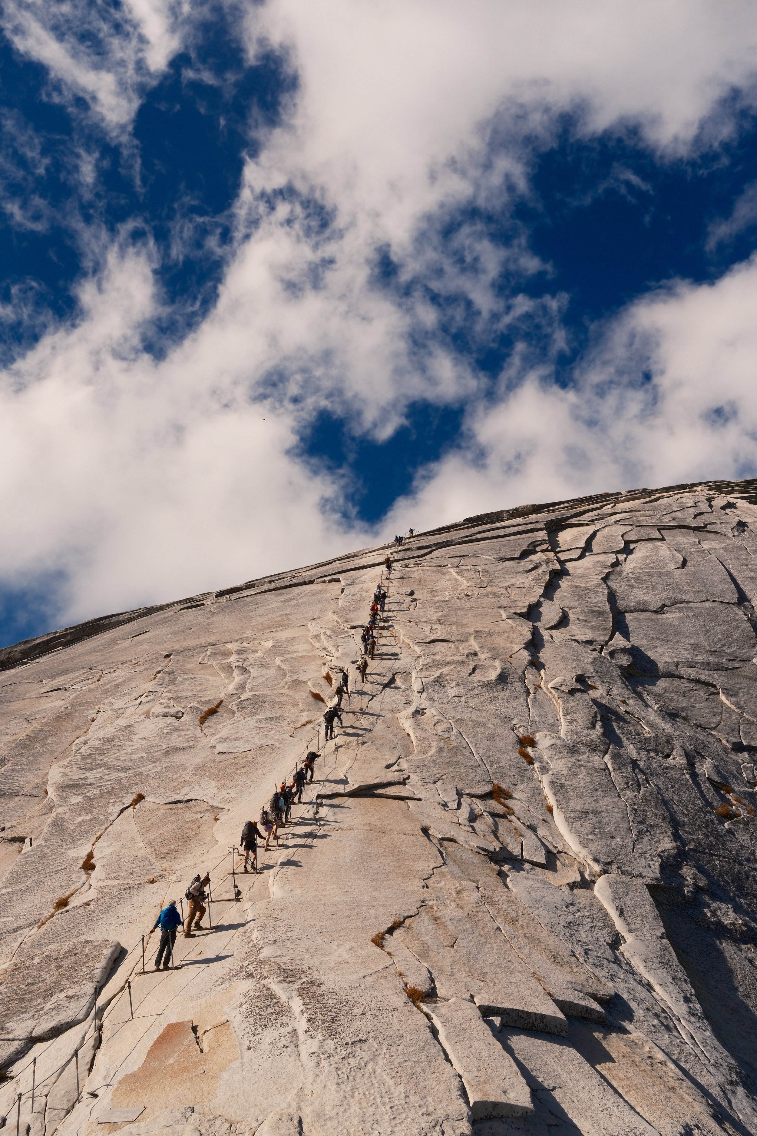 Line of hikers ascending the Half Dome cables under billowing clouds, Yosemite National Park, California, by Zyan Sachs
