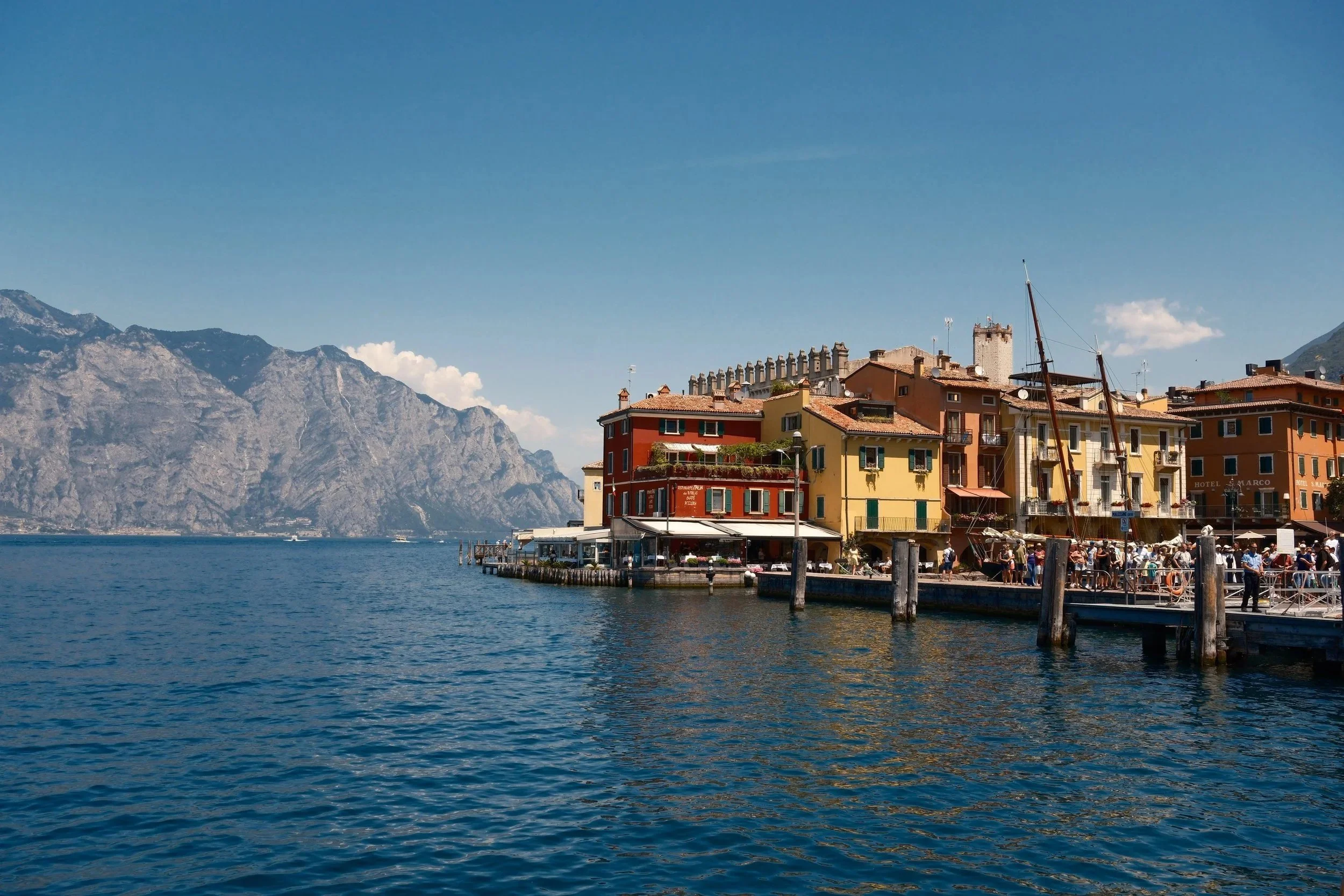 Colorful harborfront of Malcesine with its castle tower and mountains across the lake, Lake Garda, Italy, by Zyan Sachs