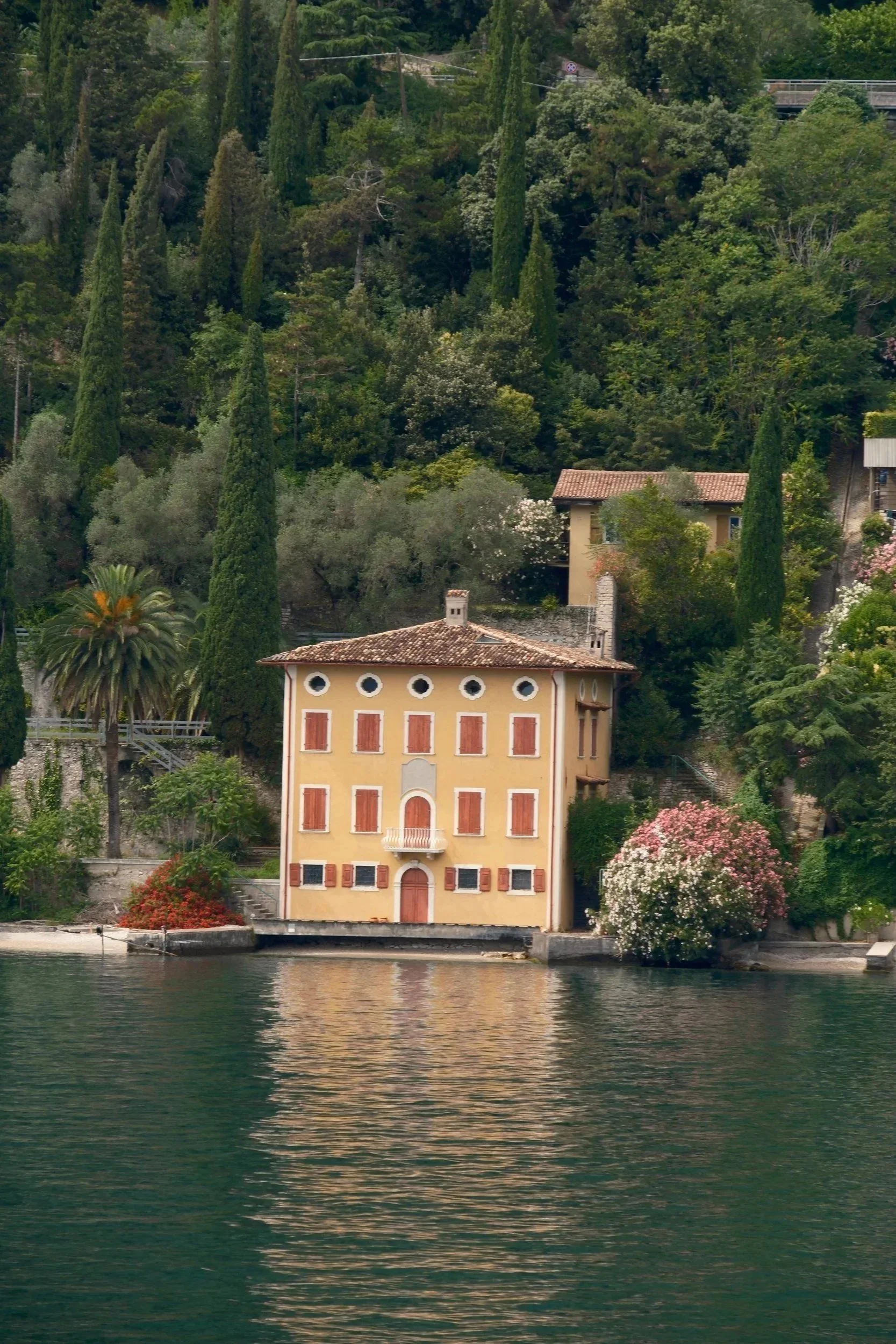 Yellow lakeside villa with red shutters among cypress trees and oleanders, reflected in green water, Lake Garda, Italy, by Zyan Sachs