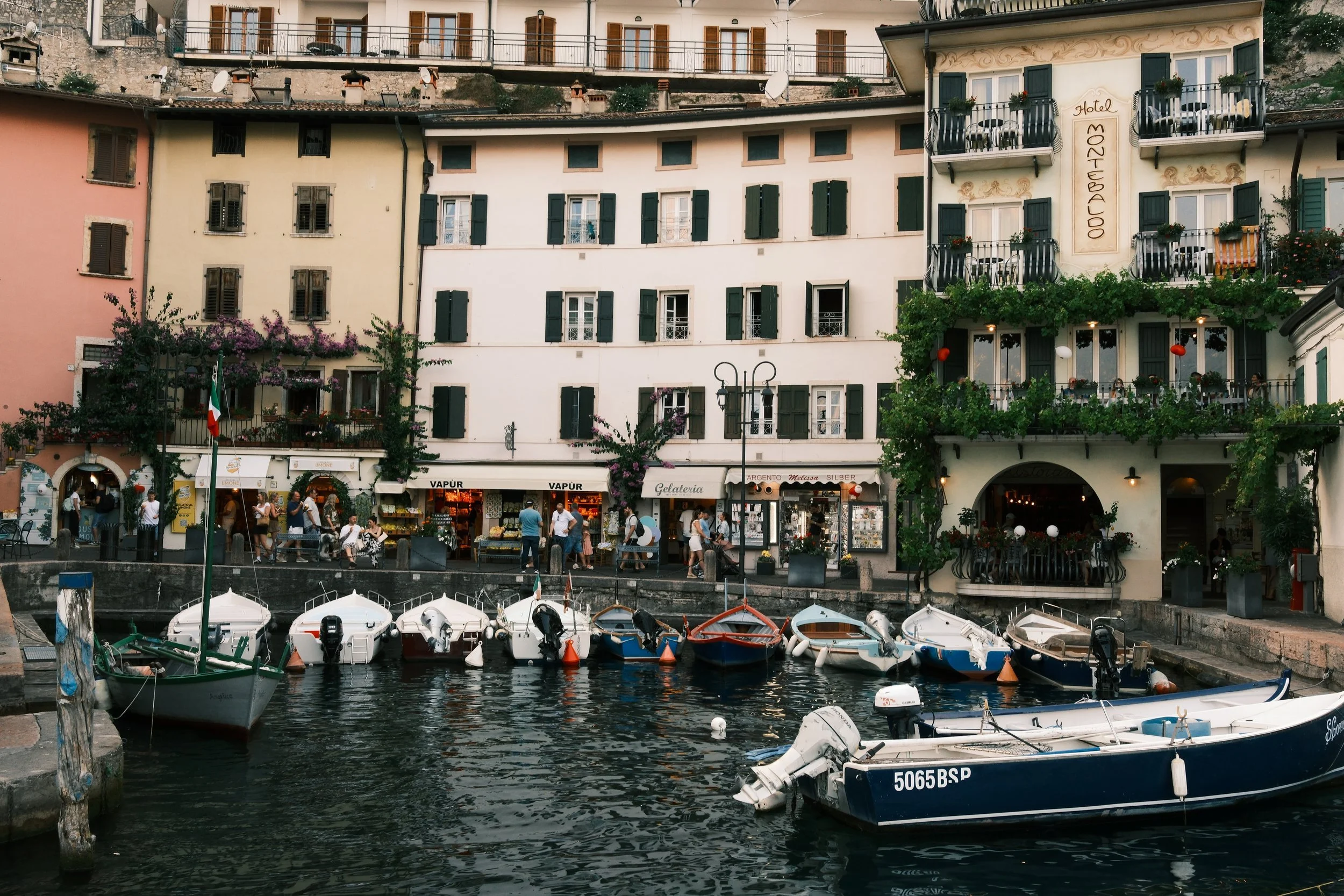 Wooden boats moored in the old harbor of Limone sul Garda below pastel buildings and the Hotel Monte Baldo, Lake Garda, Italy, by Zyan Sachs