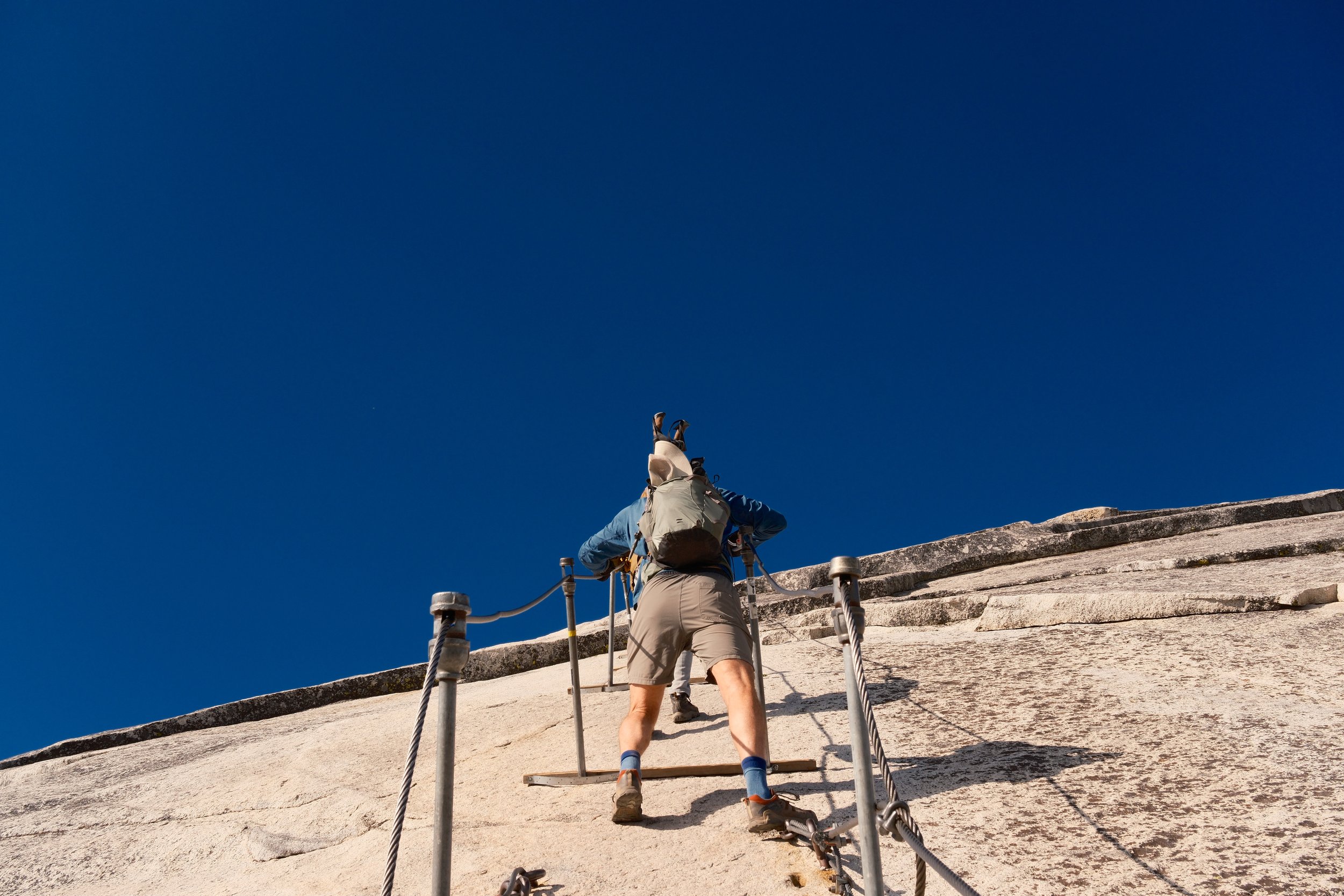 Hiker climbing the Half Dome cables against a deep blue sky, Yosemite National Park, California, by Zyan Sachs