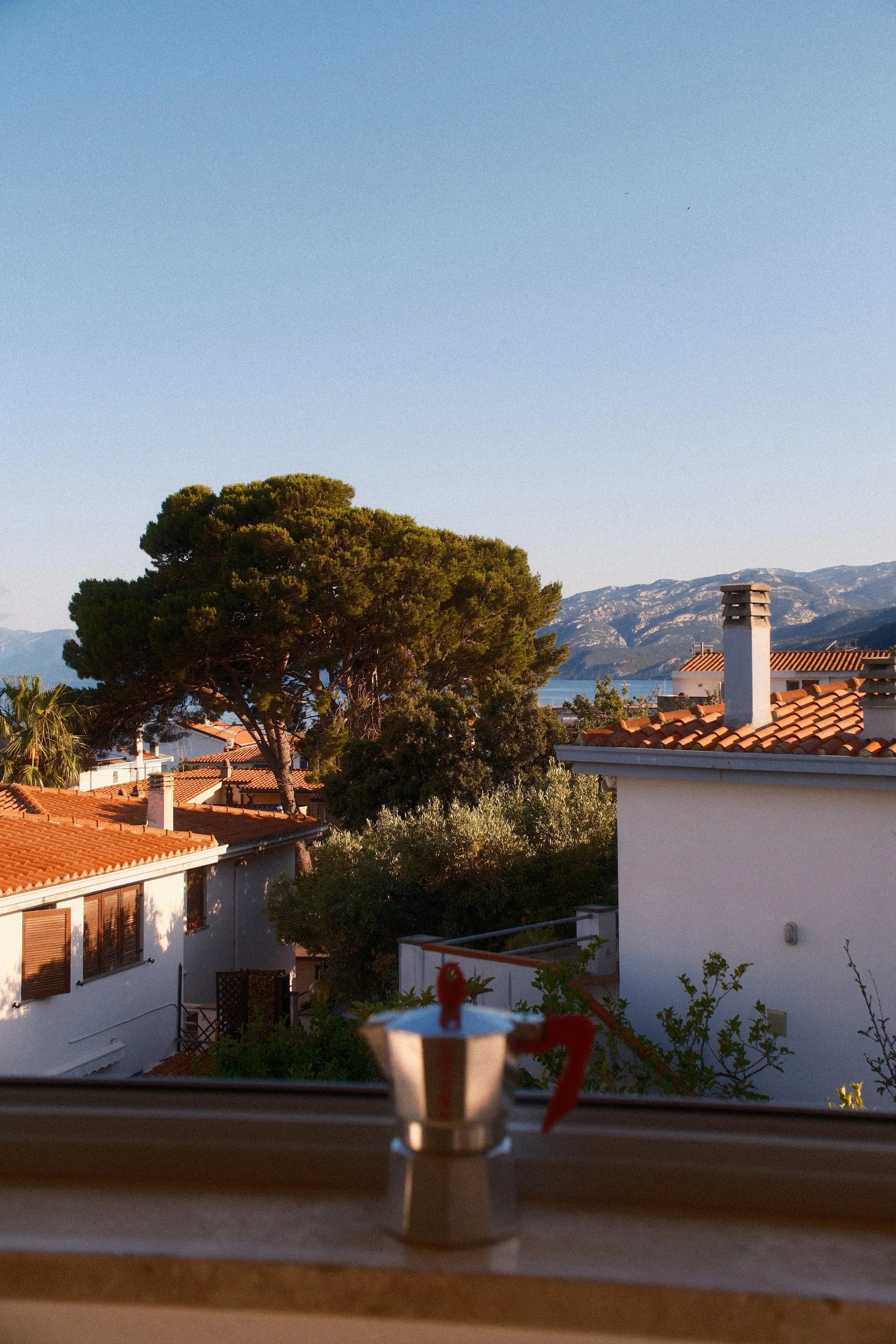 Moka pot on a sunny balcony table overlooking terracotta rooftops, pines and mountains at Casa Stefano, Sardinia, by Zyan Sachs