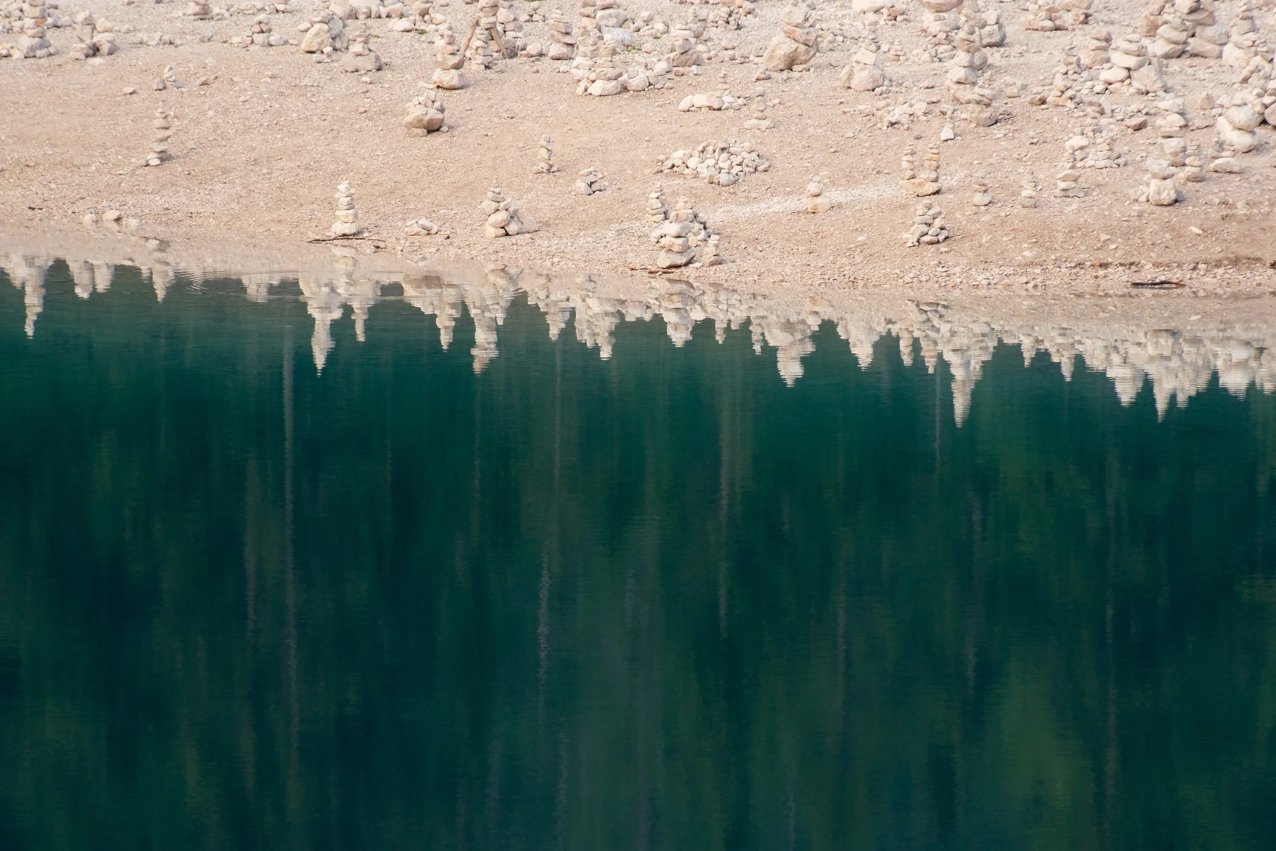 Stone cairns on the pale shore reflected in the deep green water of Lago di Braies, Dolomites, Italy, by Zyan Sachs