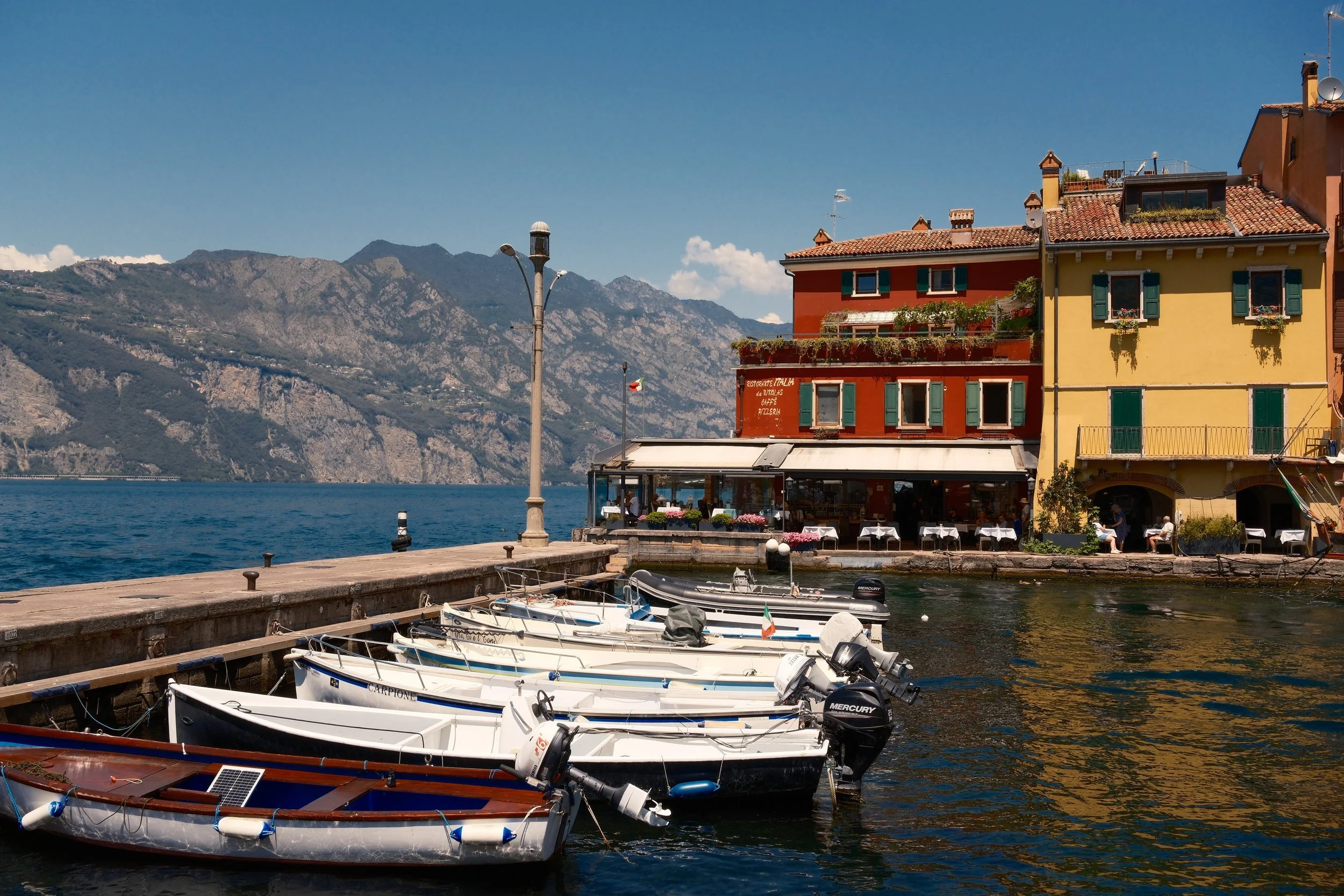 White boats moored by the pier in front of red and yellow buildings in Malcesine, Lake Garda, Italy, by Zyan Sachs