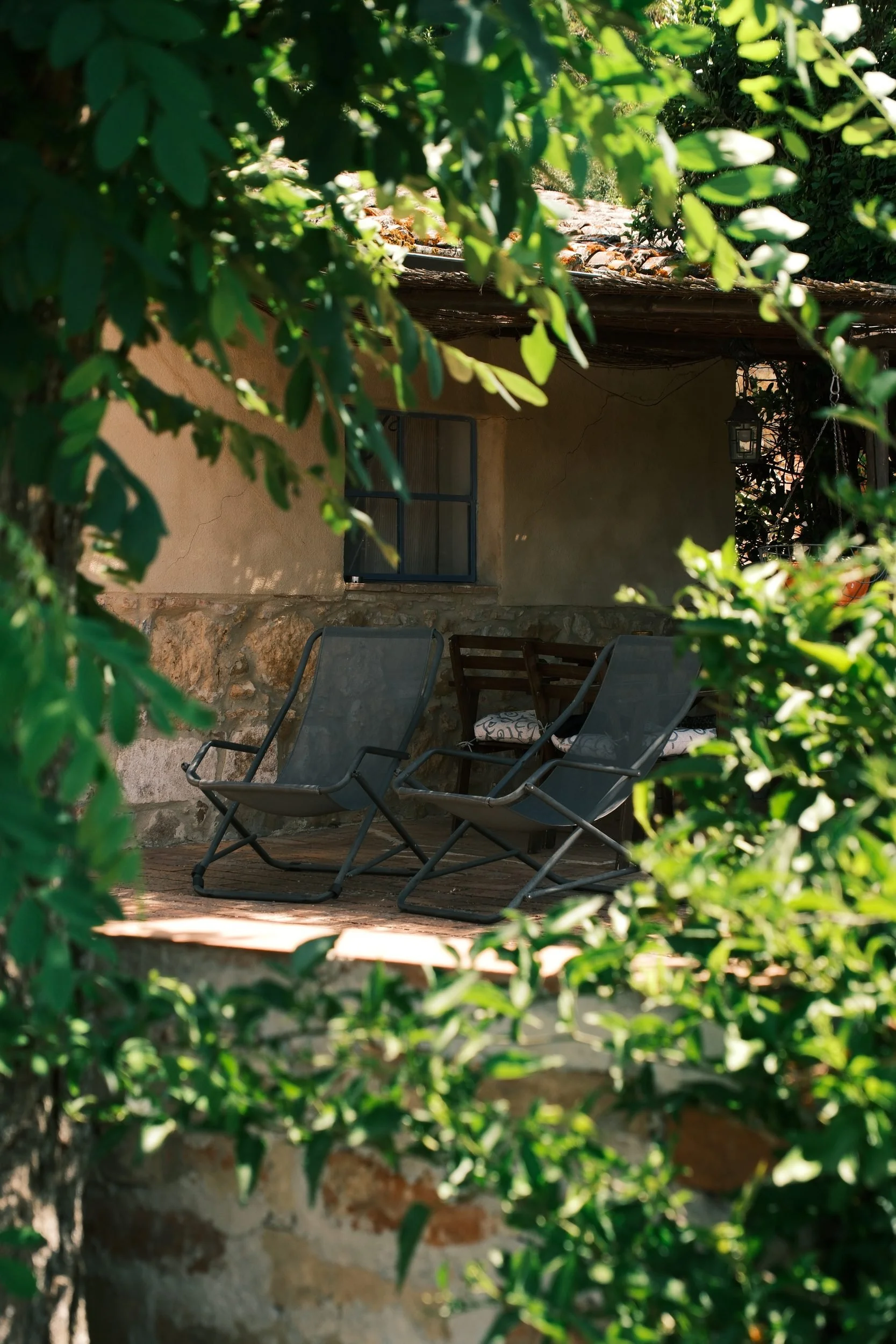 Two lounge chairs on a shaded stone farmhouse terrace seen through green leaves, Tuscany, Italy, by Zyan Sachs