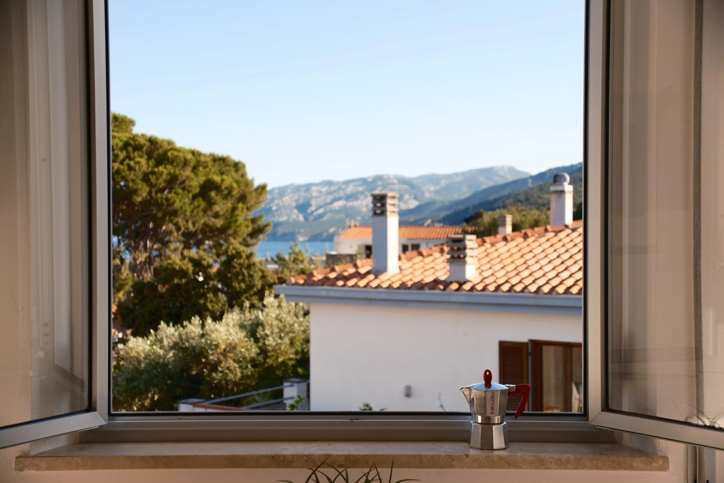 Open window with a moka pot on the sill framing rooftops, a pine tree and mountains, Sardinia, Italy, by Zyan Sachs