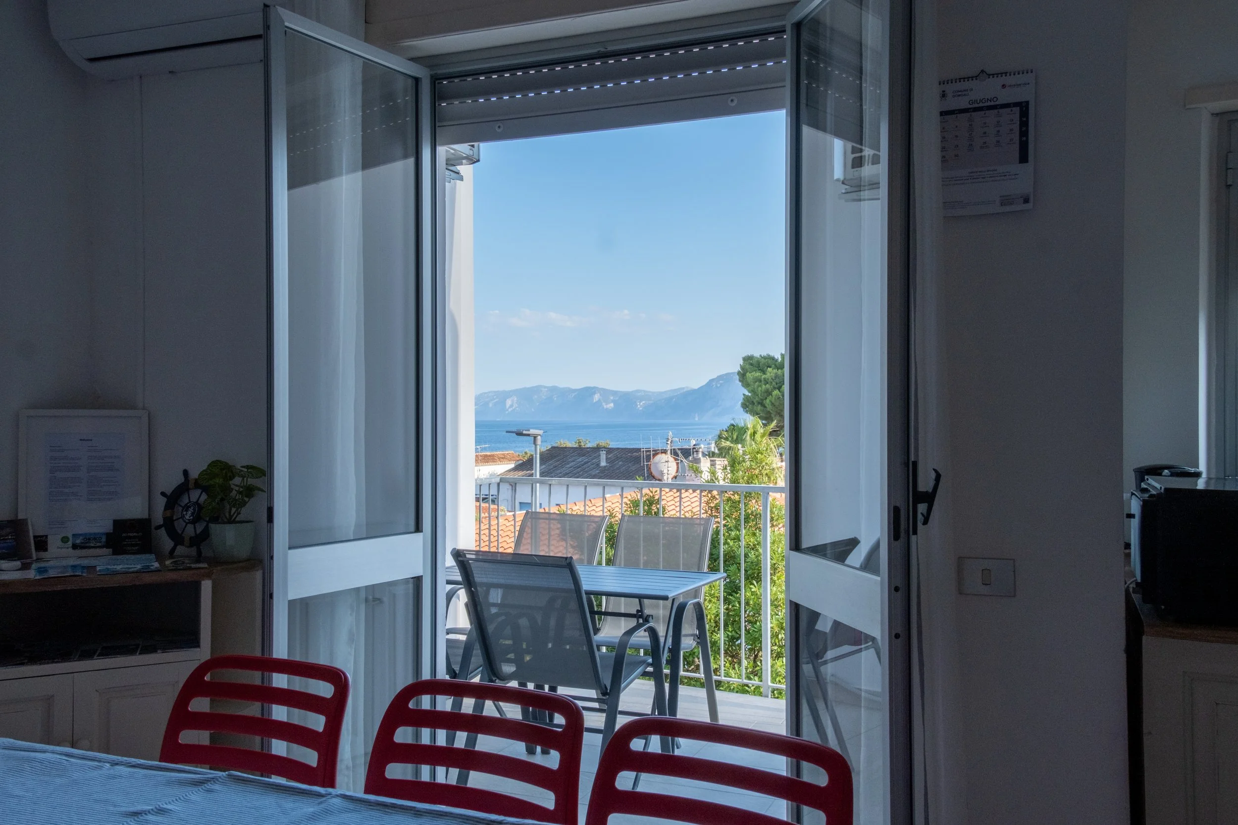 Dining table with red chairs beside open balcony doors and a sea view at Casa Stefano, Sardinia, by Zyan Sachs
