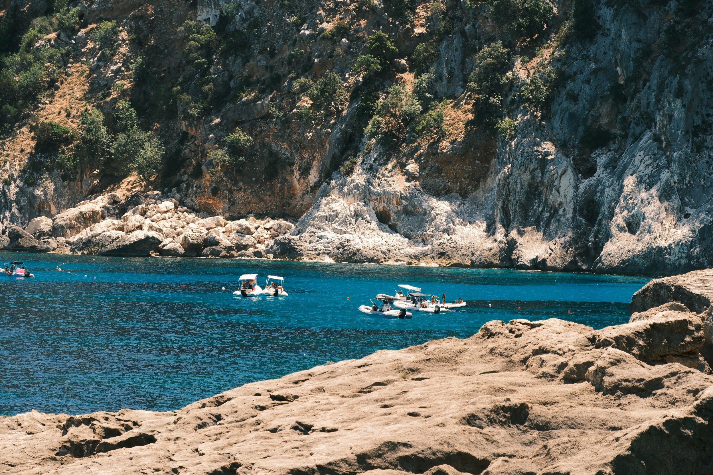 Small boats anchored in a deep blue cove beneath a limestone cliff, Gulf of Orosei, Sardinia, Italy, by Zyan Sachs
