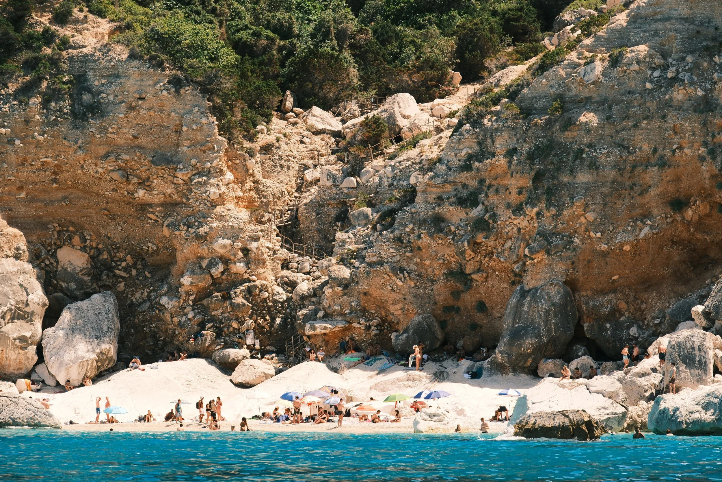 Beachgoers and colorful umbrellas on a white pebble beach below cliffs and wooden stairs, Sardinia, Italy, by Zyan Sachs
