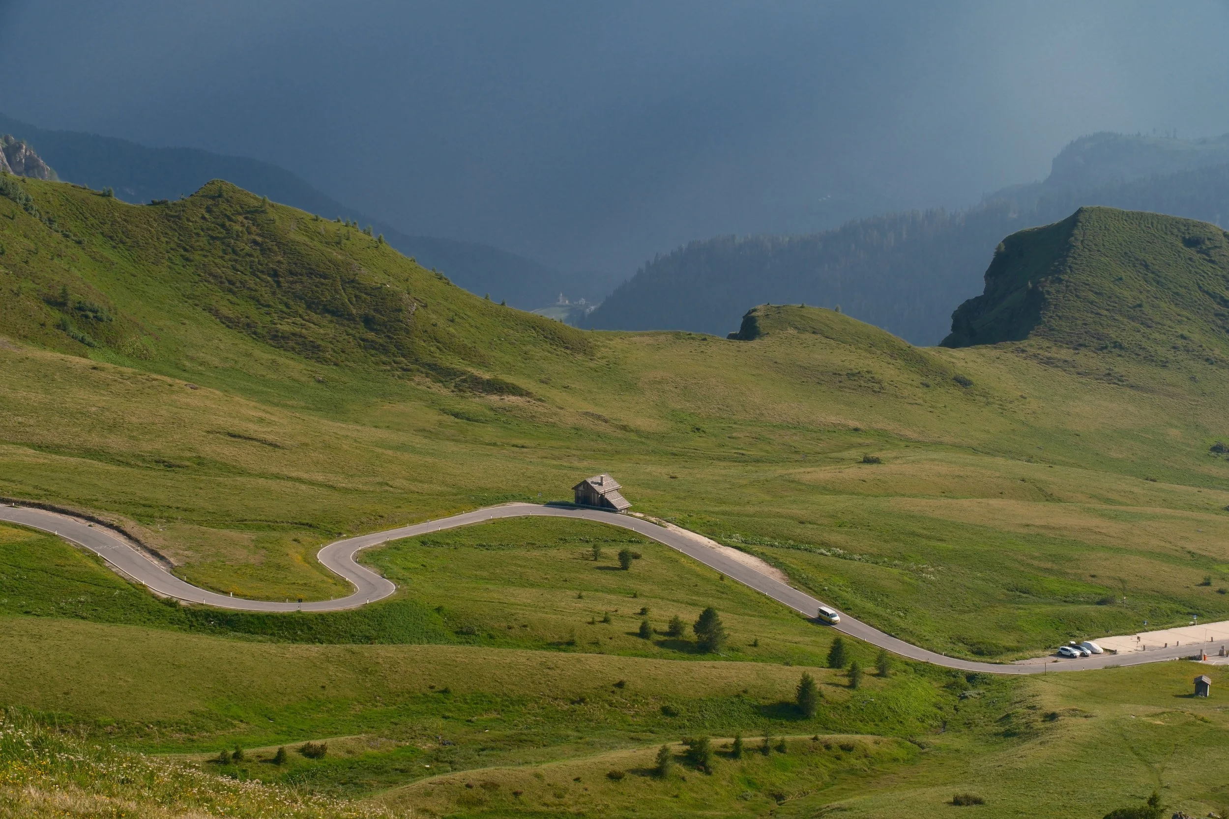 Winding mountain road and lone hut across green alpine meadows under a dark storm sky near Passo Giau, Dolomites, Italy, by Zyan Sachs