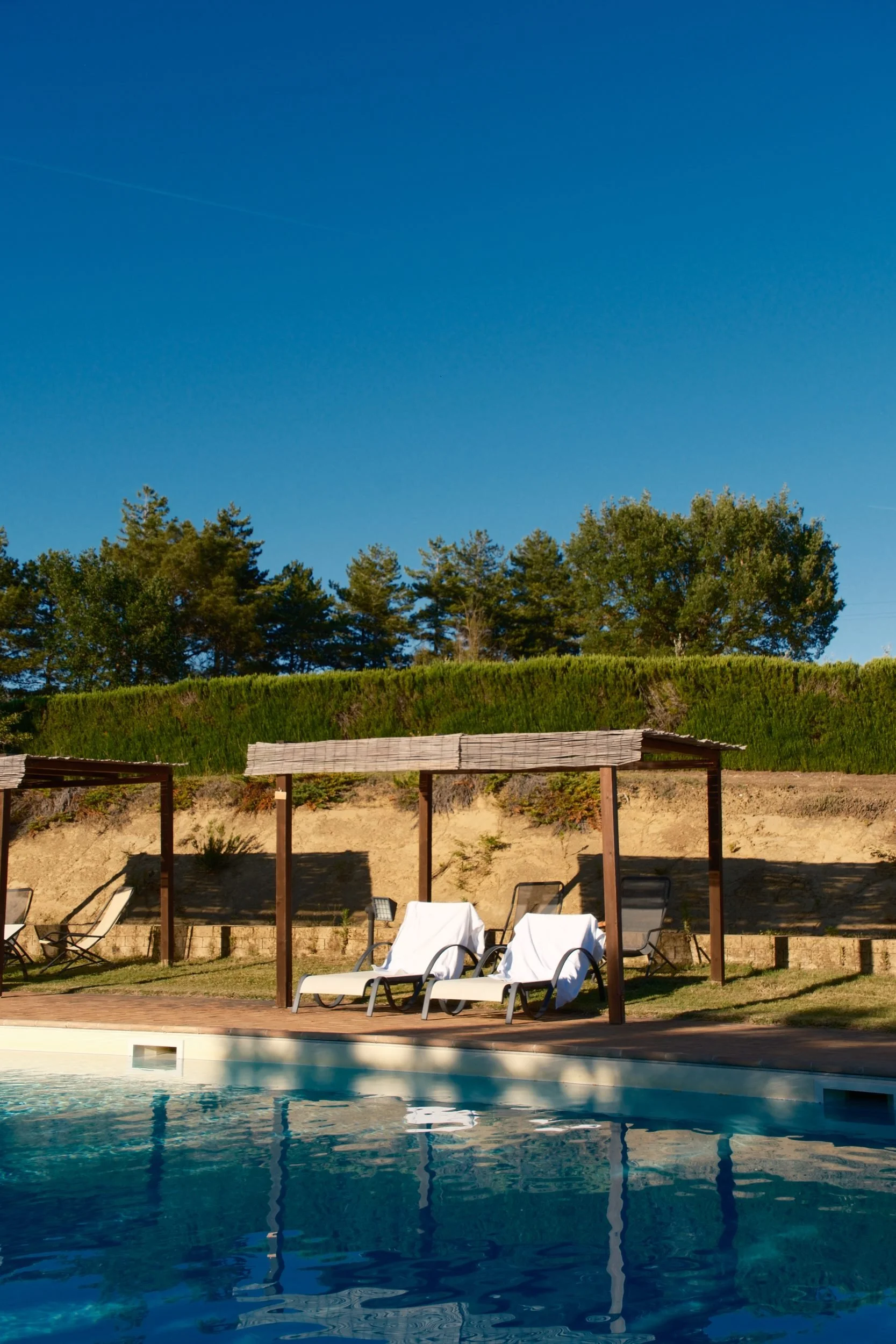Pool with white loungers under wooden pergolas against a deep blue sky, Tuscan farmhouse stay, Italy, by Zyan Sachs