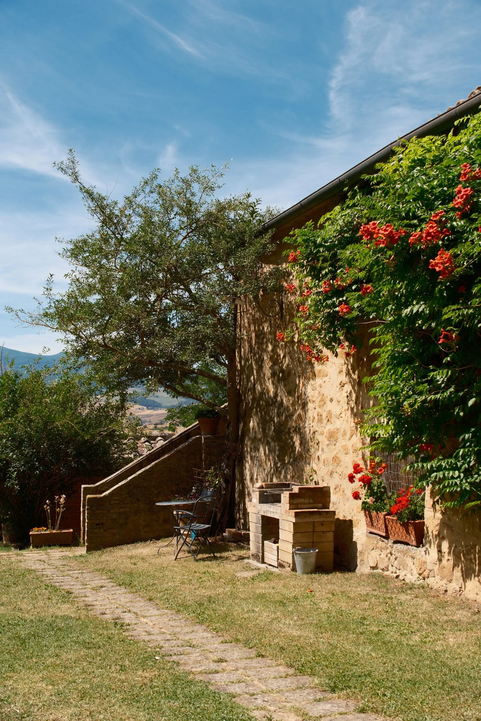 Stone farmhouse wall covered in orange trumpet vine flowers with a garden path and brick grill, Tuscany, Italy, by Zyan Sachs