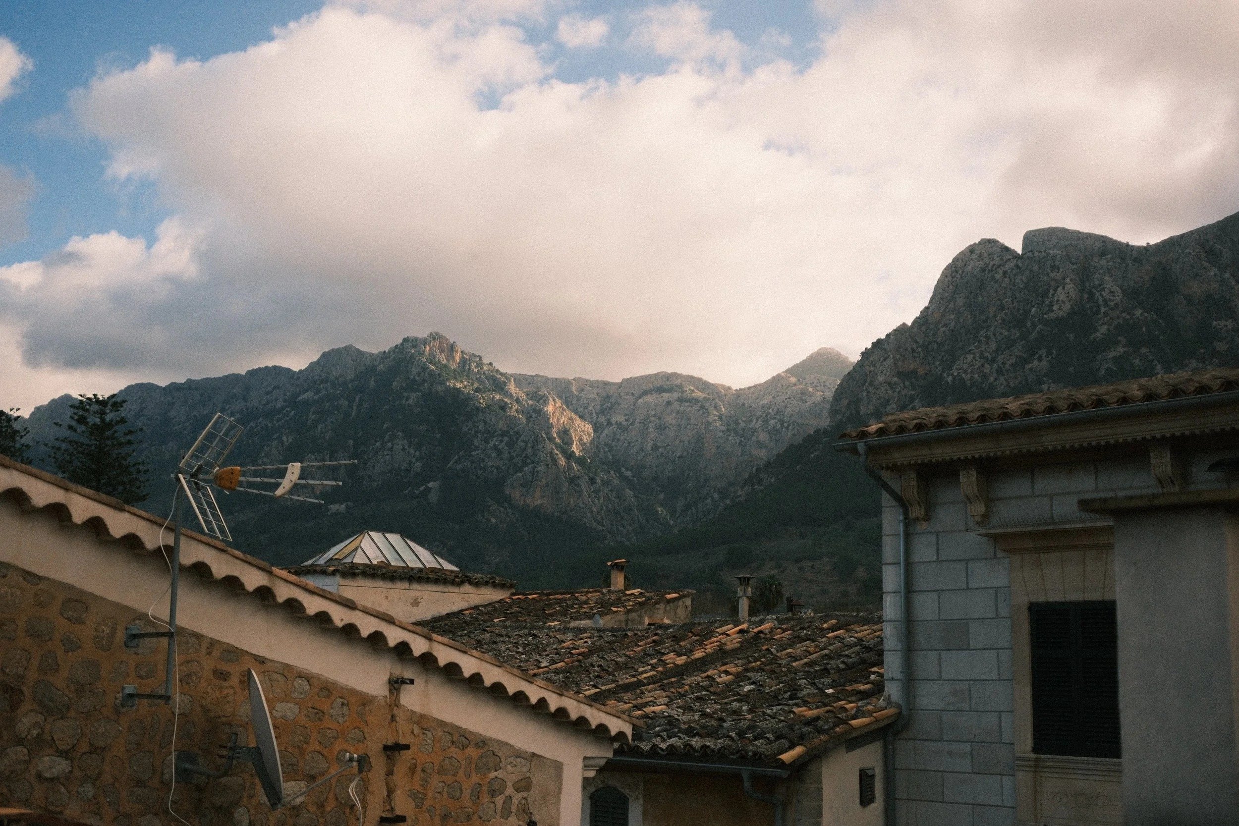 Stone village rooftops below the Tramuntana mountains under billowing clouds, Mallorca, Spain - fine art print by Zyan Sachs