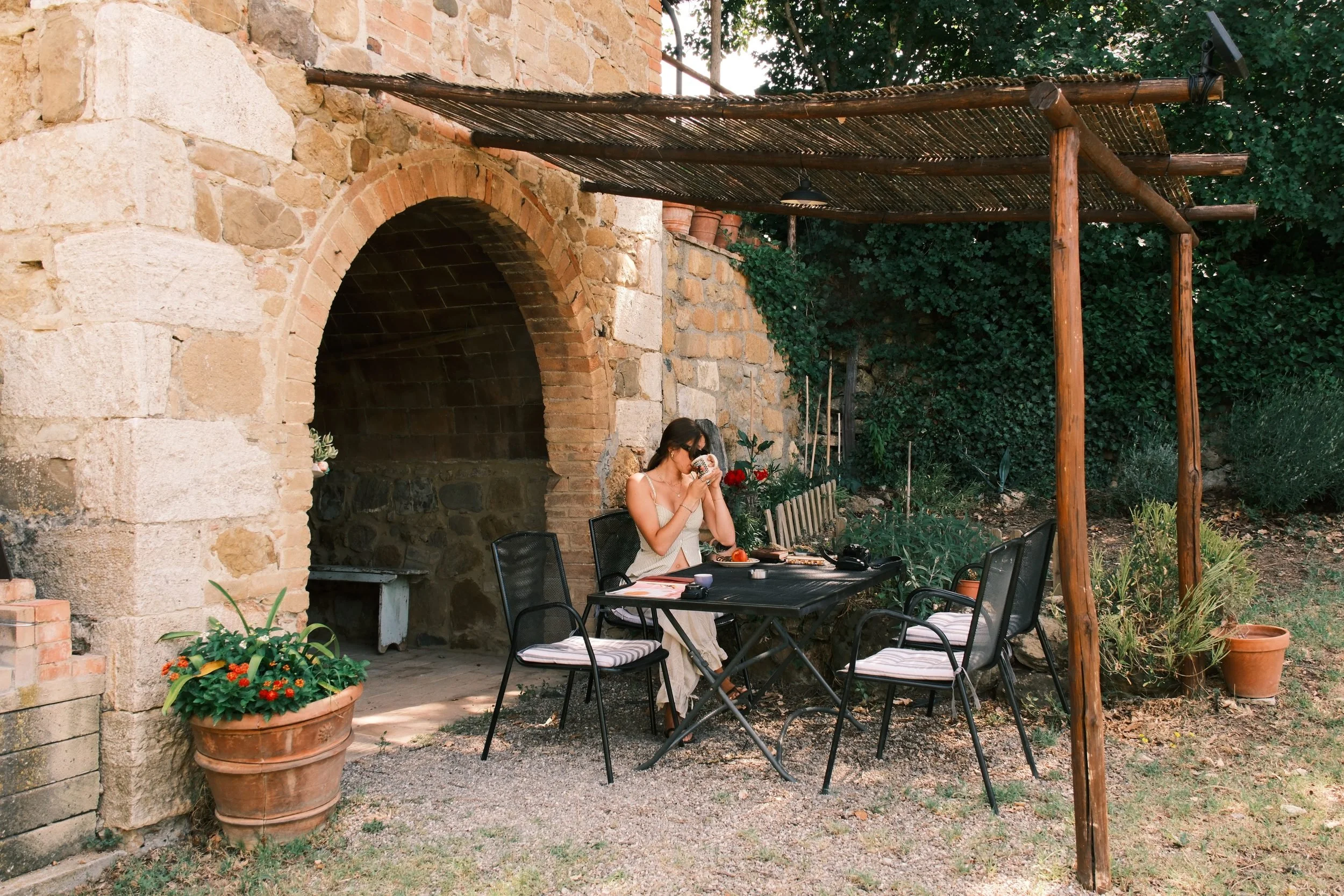 Woman having breakfast at a table under a wooden pergola beside a stone arch, Tuscan farmhouse, Italy, by Zyan Sachs