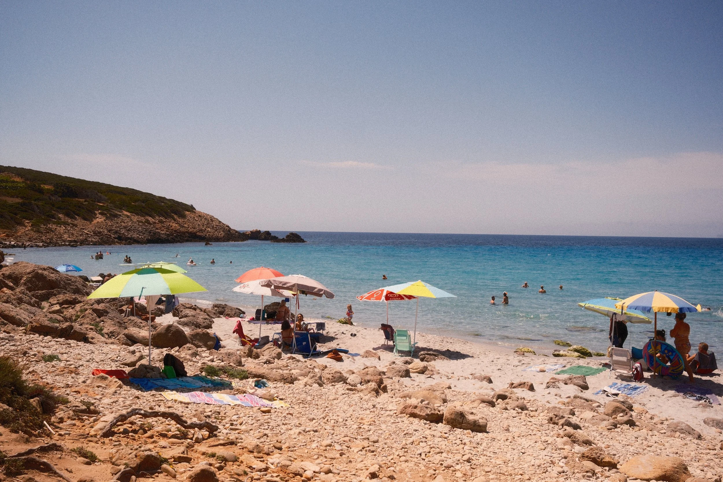 Colorful beach umbrellas on a rocky beach along a turquoise bay, Sardinia, Italy, by Zyan Sachs