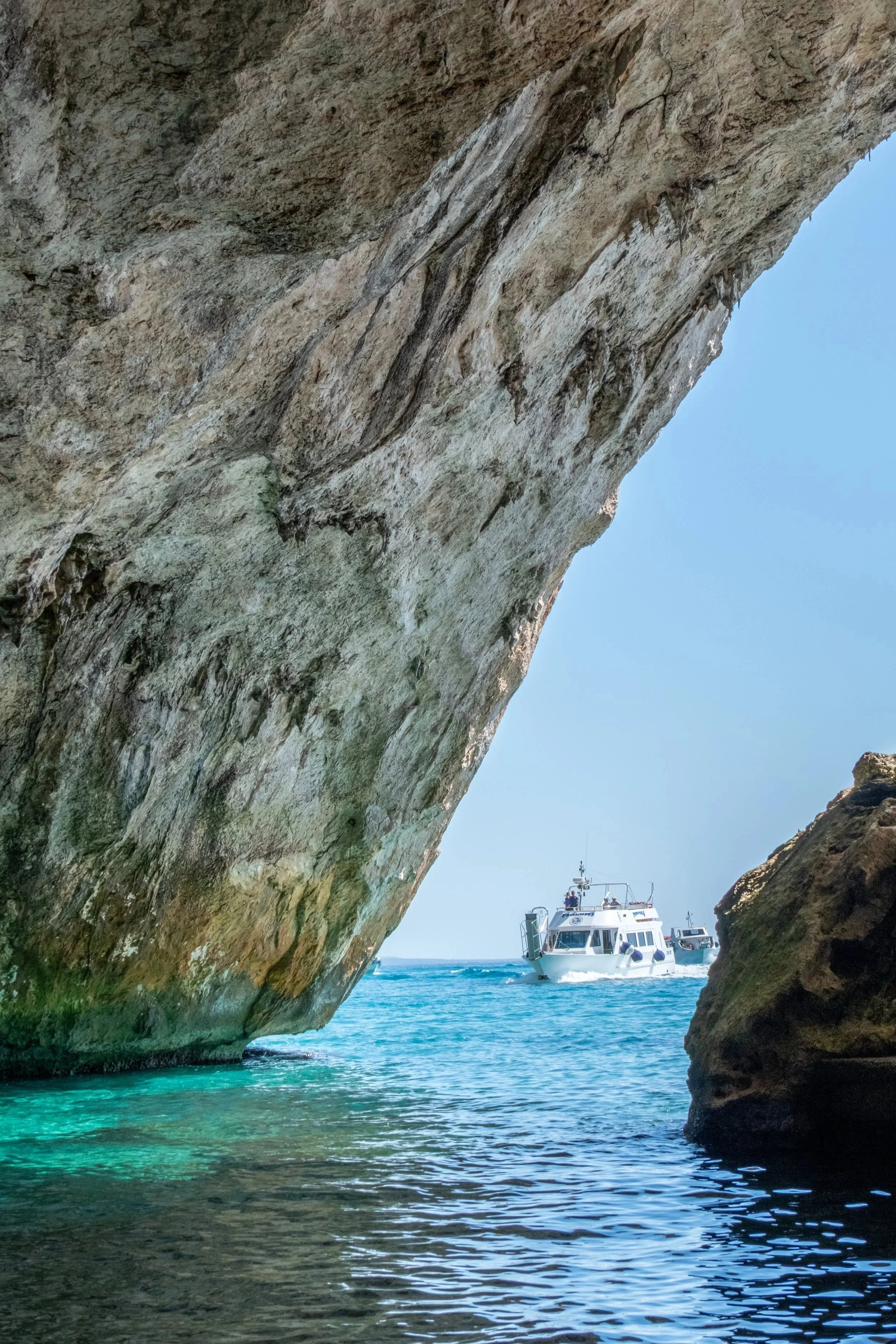 Boat passing a limestone sea arch over turquoise water near Cala Gonone, Gulf of Orosei, Sardinia, by Zyan Sachs for Gonone Rental