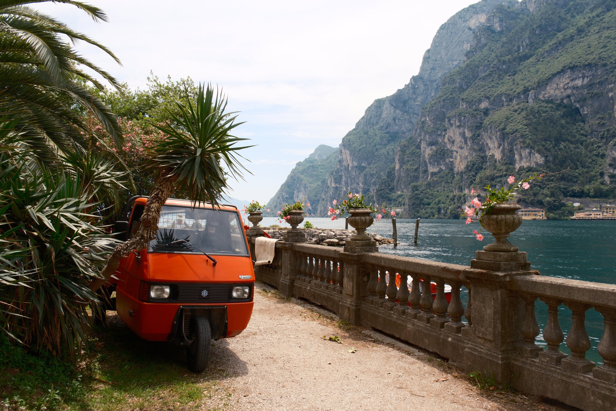 Orange Piaggio Ape parked by palms and a stone balustrade on the lakeshore near Riva del Garda, Lake Garda, Italy, by Zyan Sachs