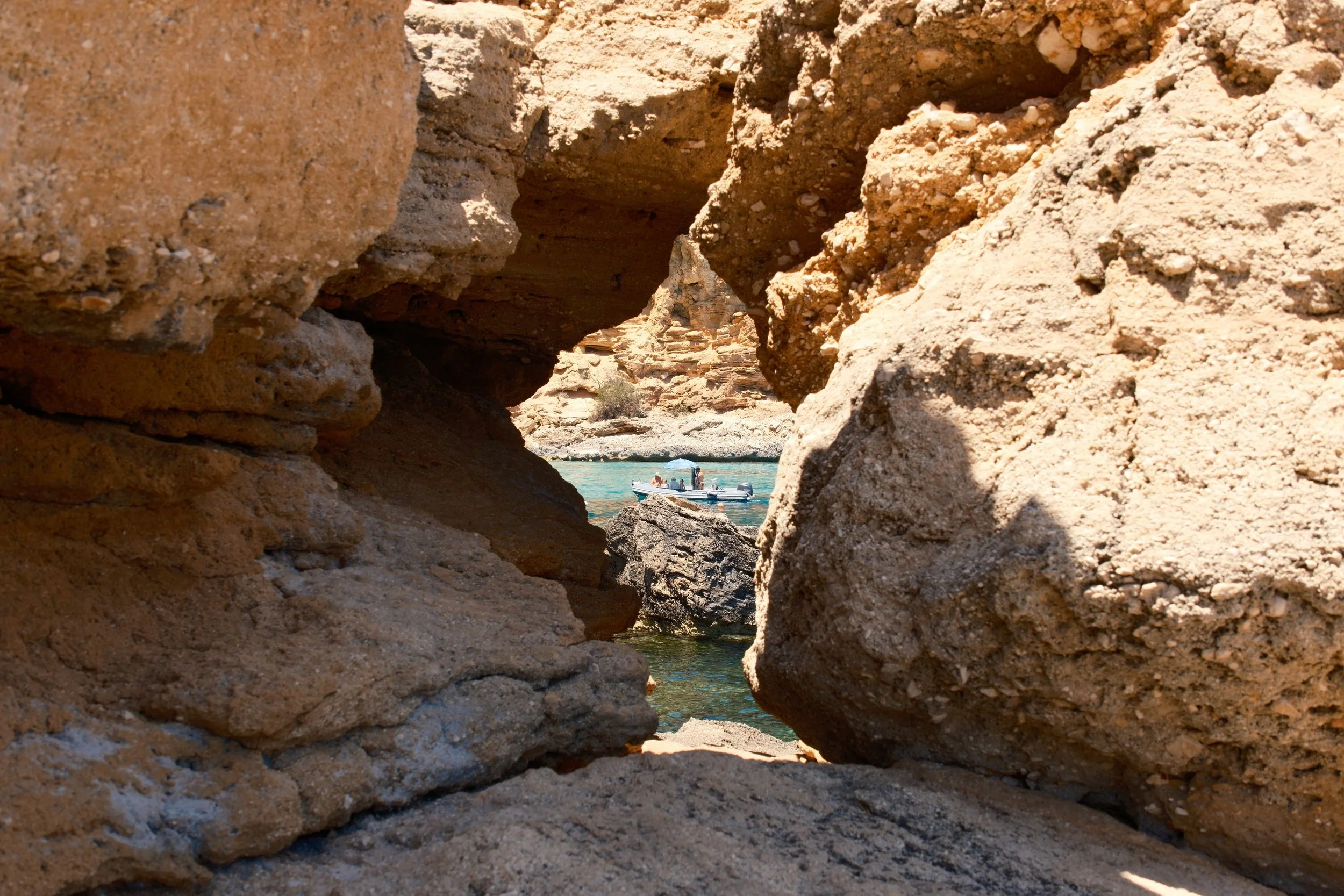 Boat on turquoise water glimpsed through a gap in golden sandstone rocks, Sardinia, Italy, by Zyan Sachs