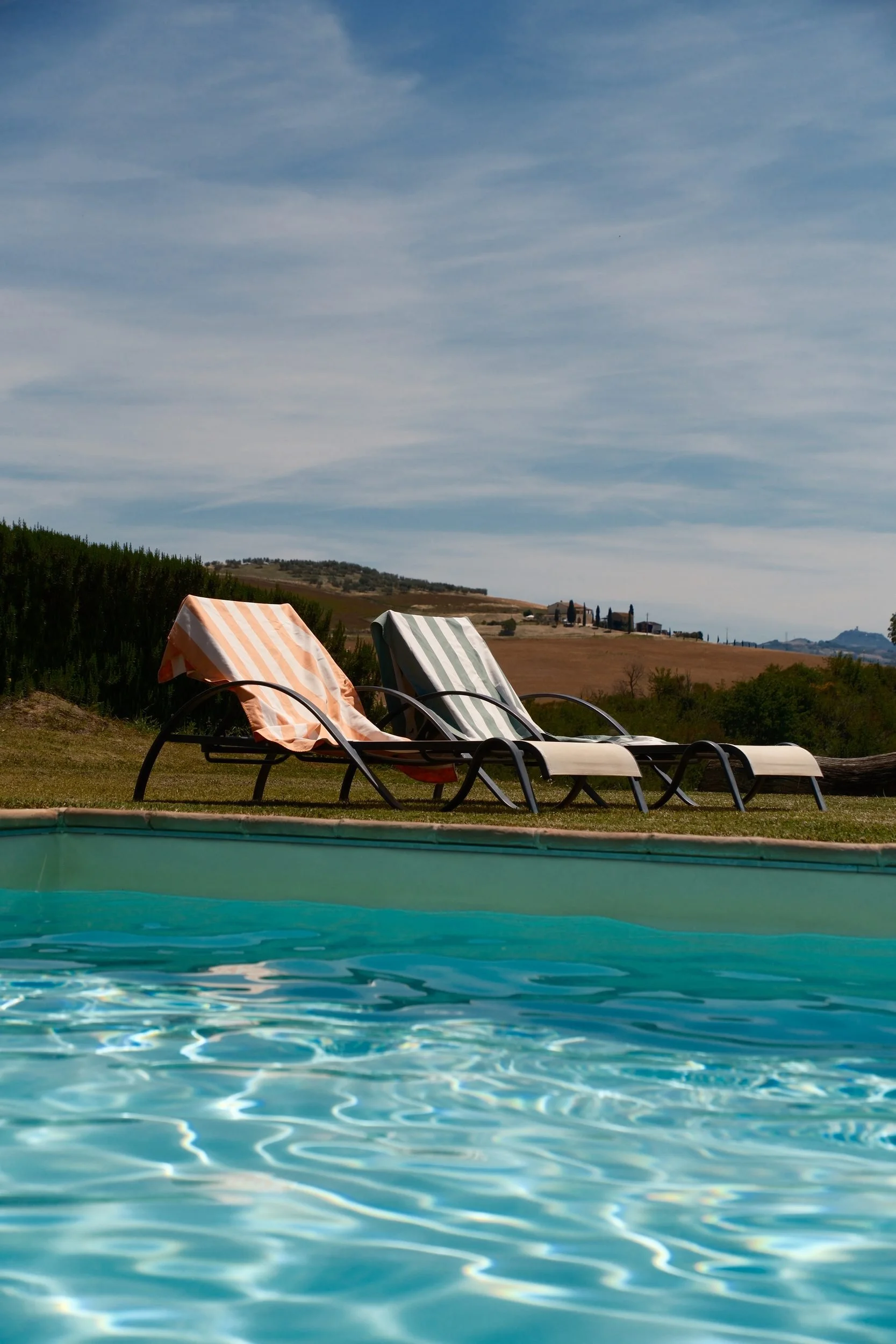 Two pool loungers with striped towels beside a turquoise pool overlooking the Tuscan countryside, Italy, by Zyan Sachs
