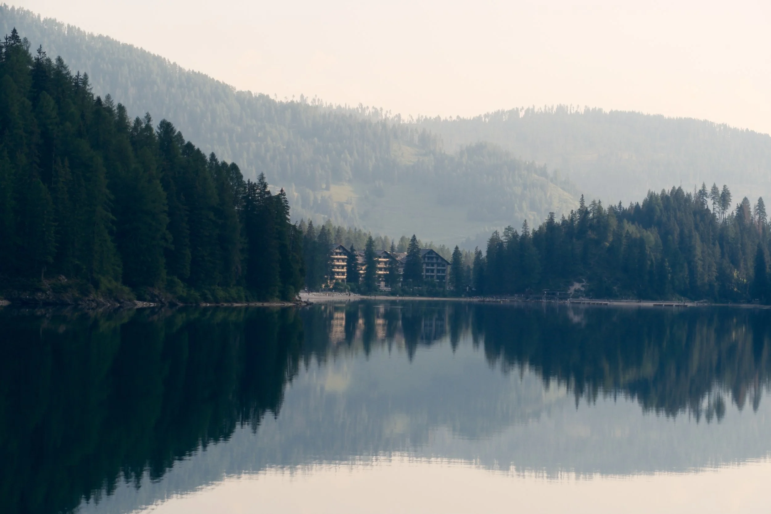 Hotel Lago di Braies among misty forested hills reflected in calm water at dawn, Dolomites, Italy, by Zyan Sachs