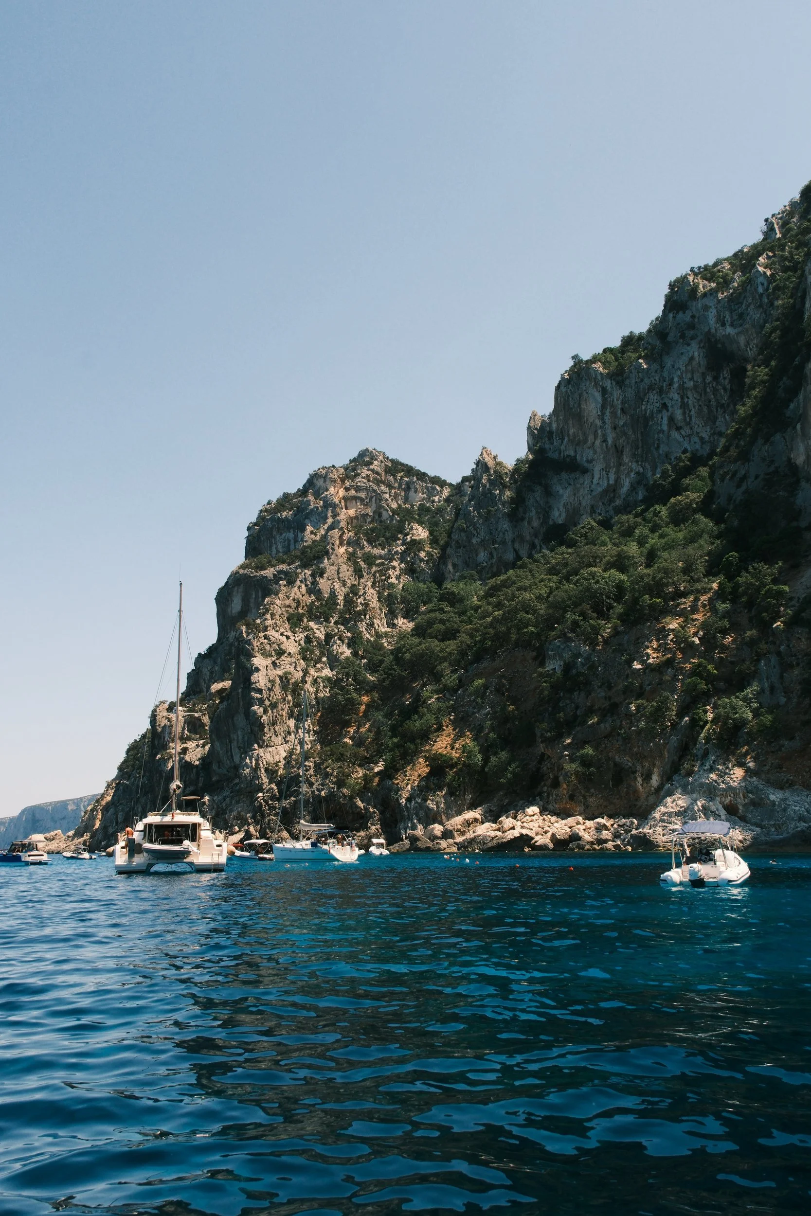 Catamaran and sailboats anchored below steep green cliffs on deep blue water, Gulf of Orosei, Sardinia, Italy, by Zyan Sachs