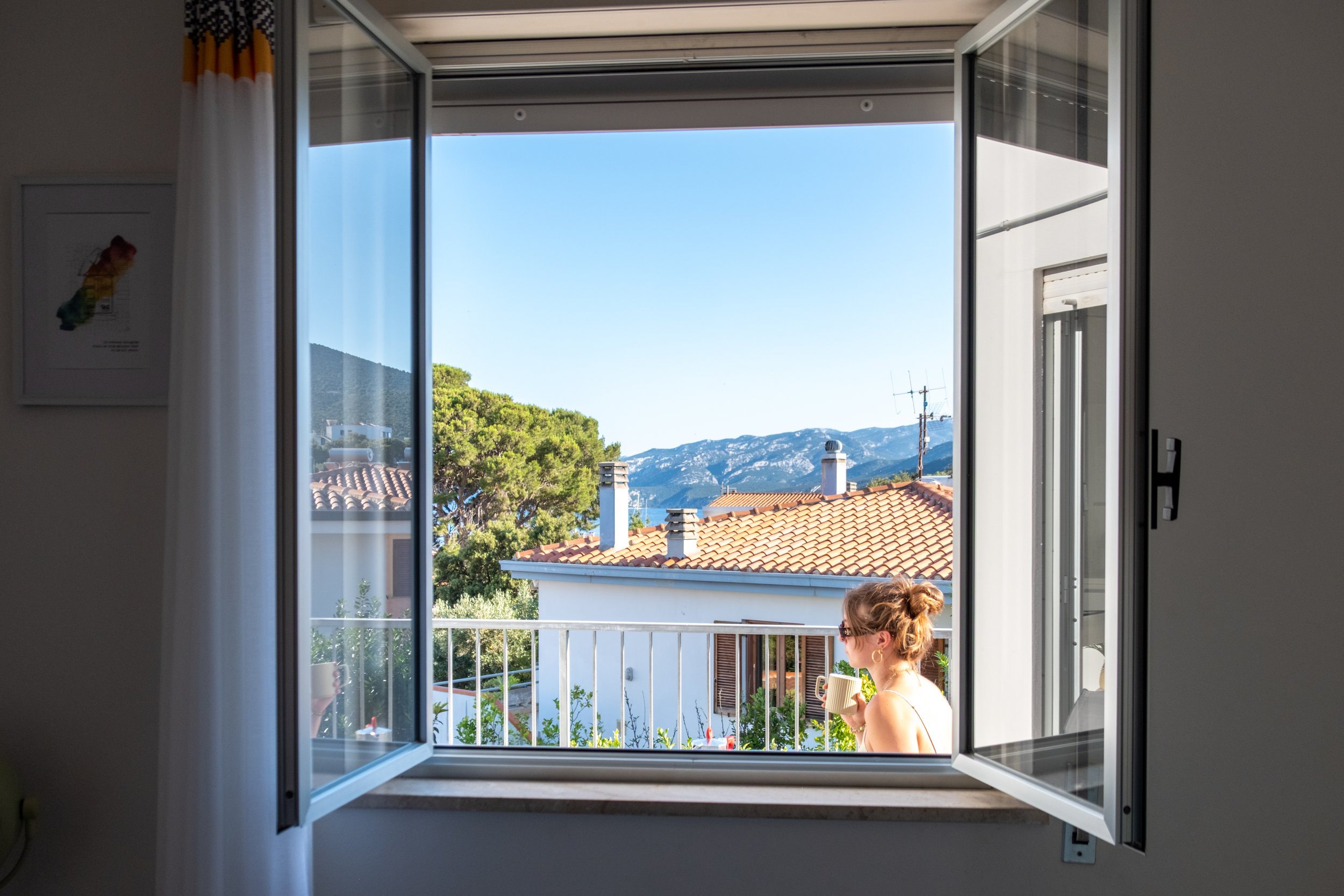 Open window framing rooftops, trees and the sea with a guest on the balcony at Casa Stefano, Sardinia, by Zyan Sachs