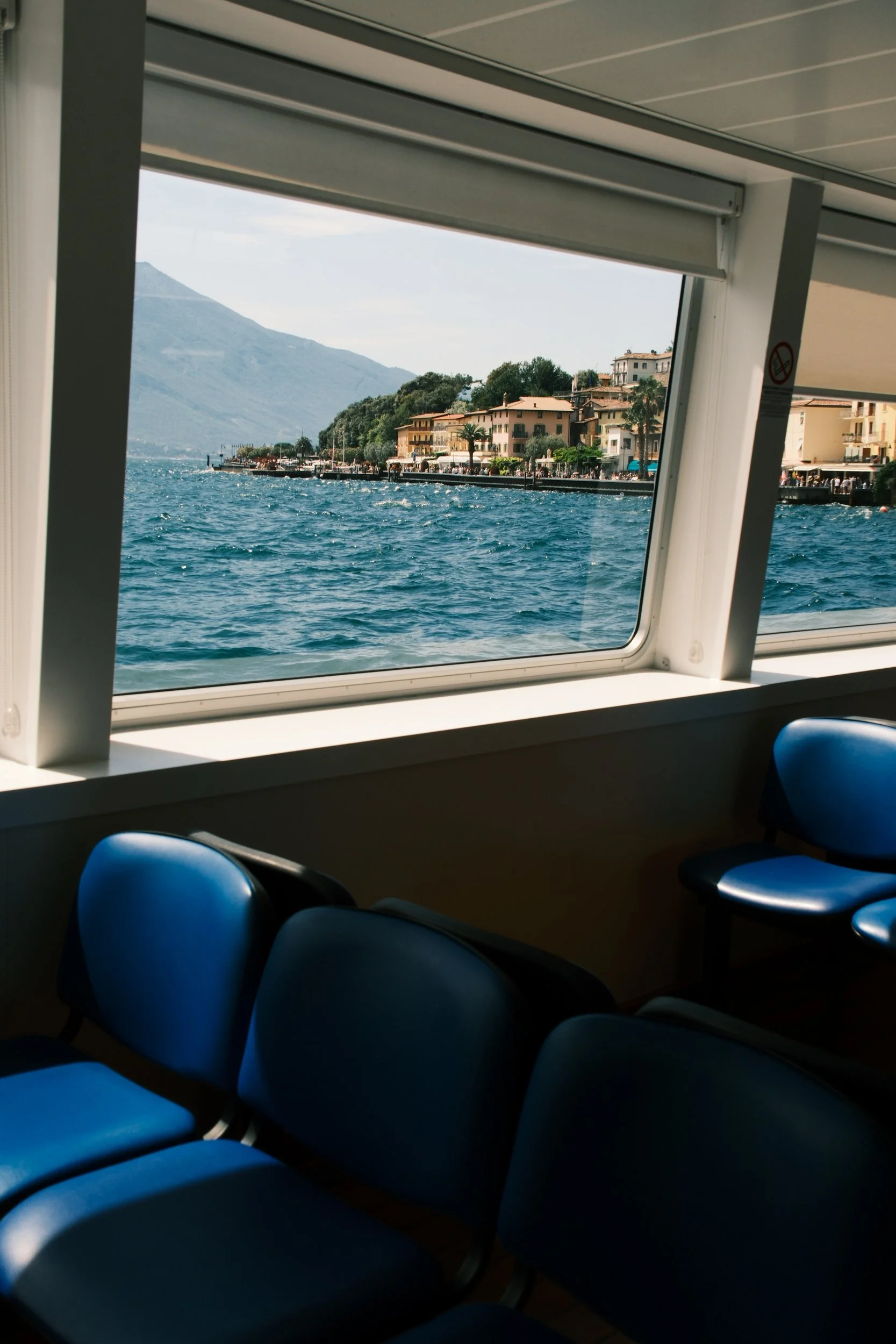 View of Limone sul Garda through a ferry window above blue seats and choppy water, Lake Garda, Italy, by Zyan Sachs
